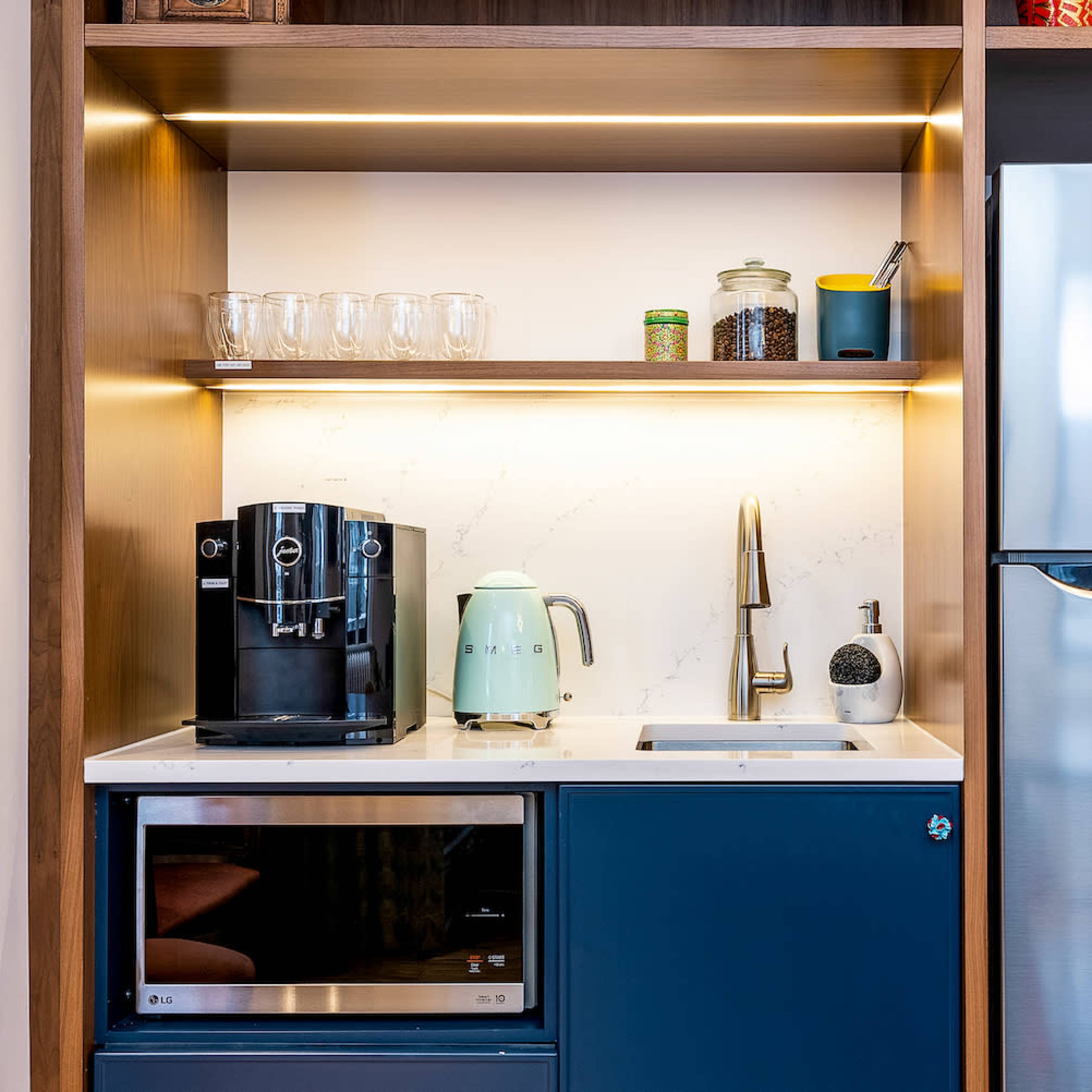 A modern kitchenette featuring a coffee maker, kettle, sink, and refrigerator, all arranged within wooden cabinetry with illuminated shelves.