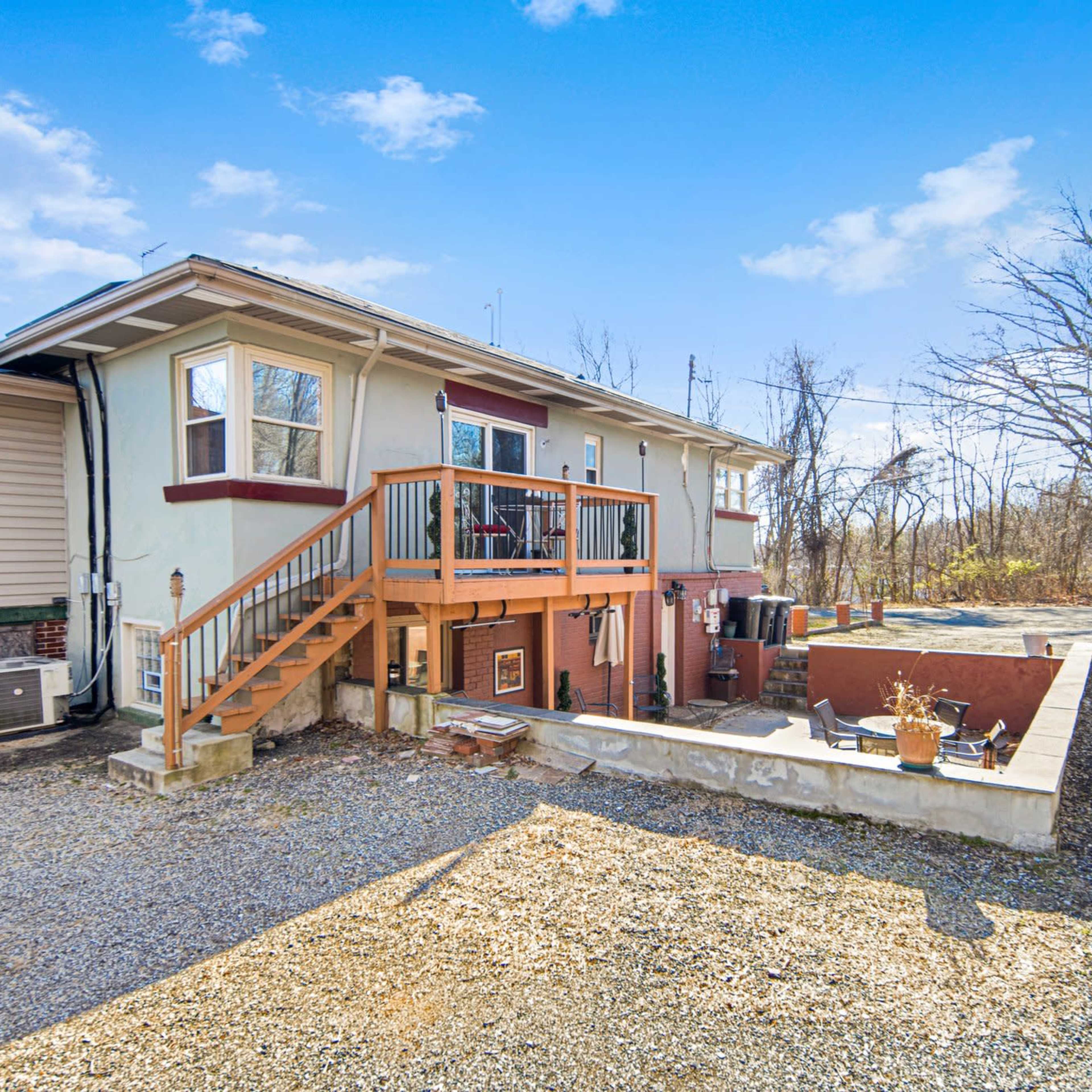 A two-story house with a wooden stairway leading to a deck on the upper level, surrounded by a gravel area and bare trees in the background.