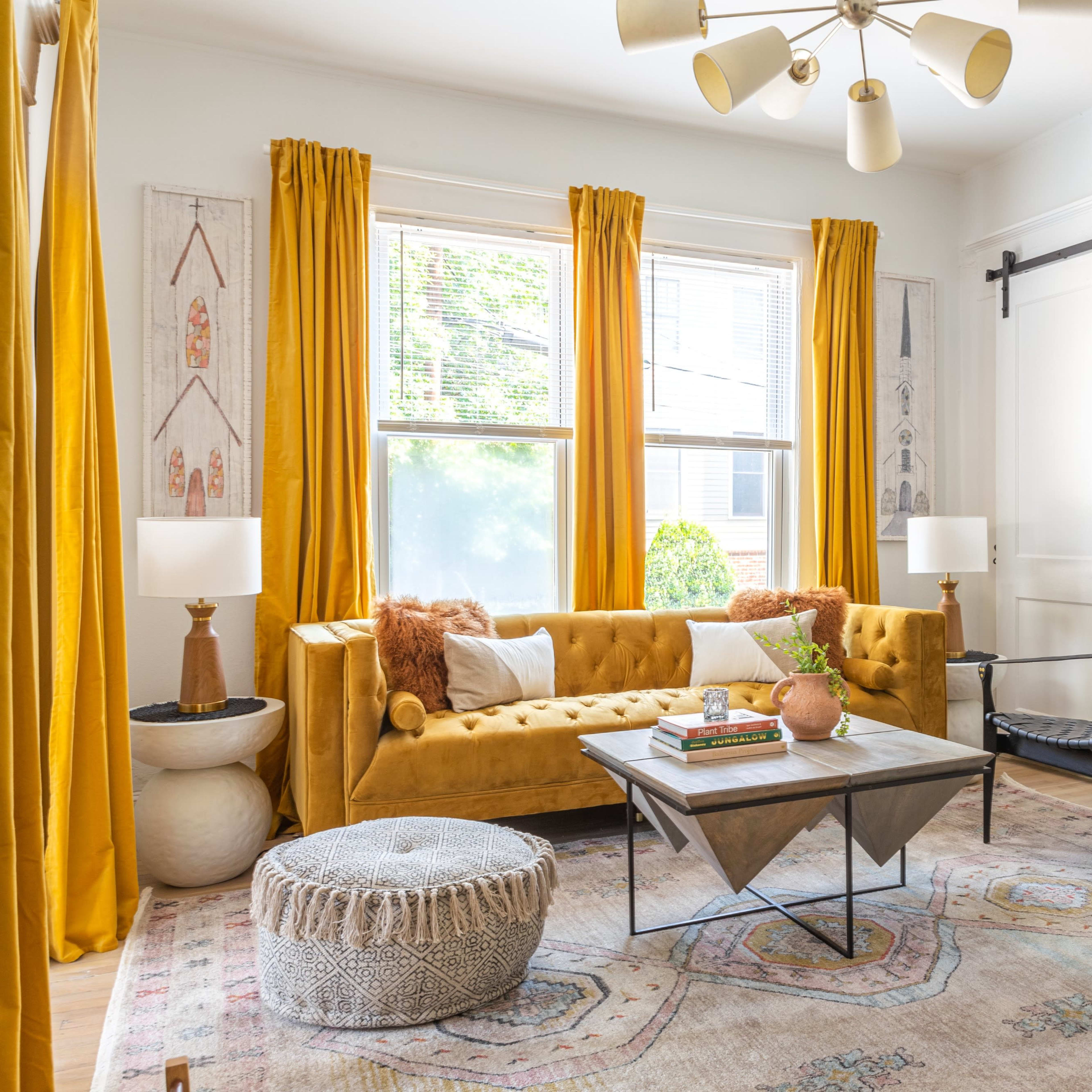 The living room features a mustard yellow sofa, a black chair, and a coffee table on a patterned rug, surrounded by large windows with yellow curtains.