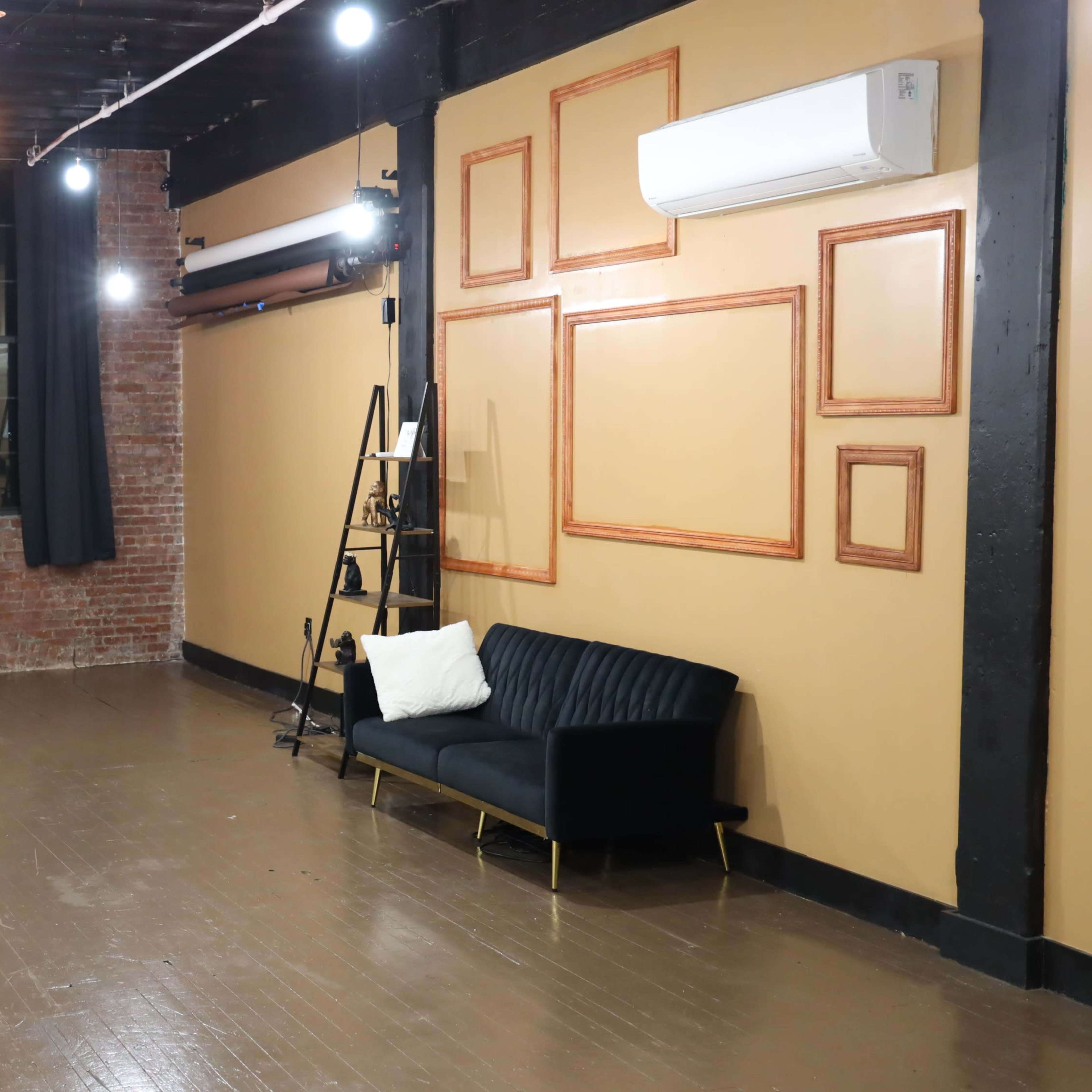 An empty room with exposed brick walls, a large window, wooden flooring, and a black sofa against one wall, illuminated by string lights and ceiling fixtures.