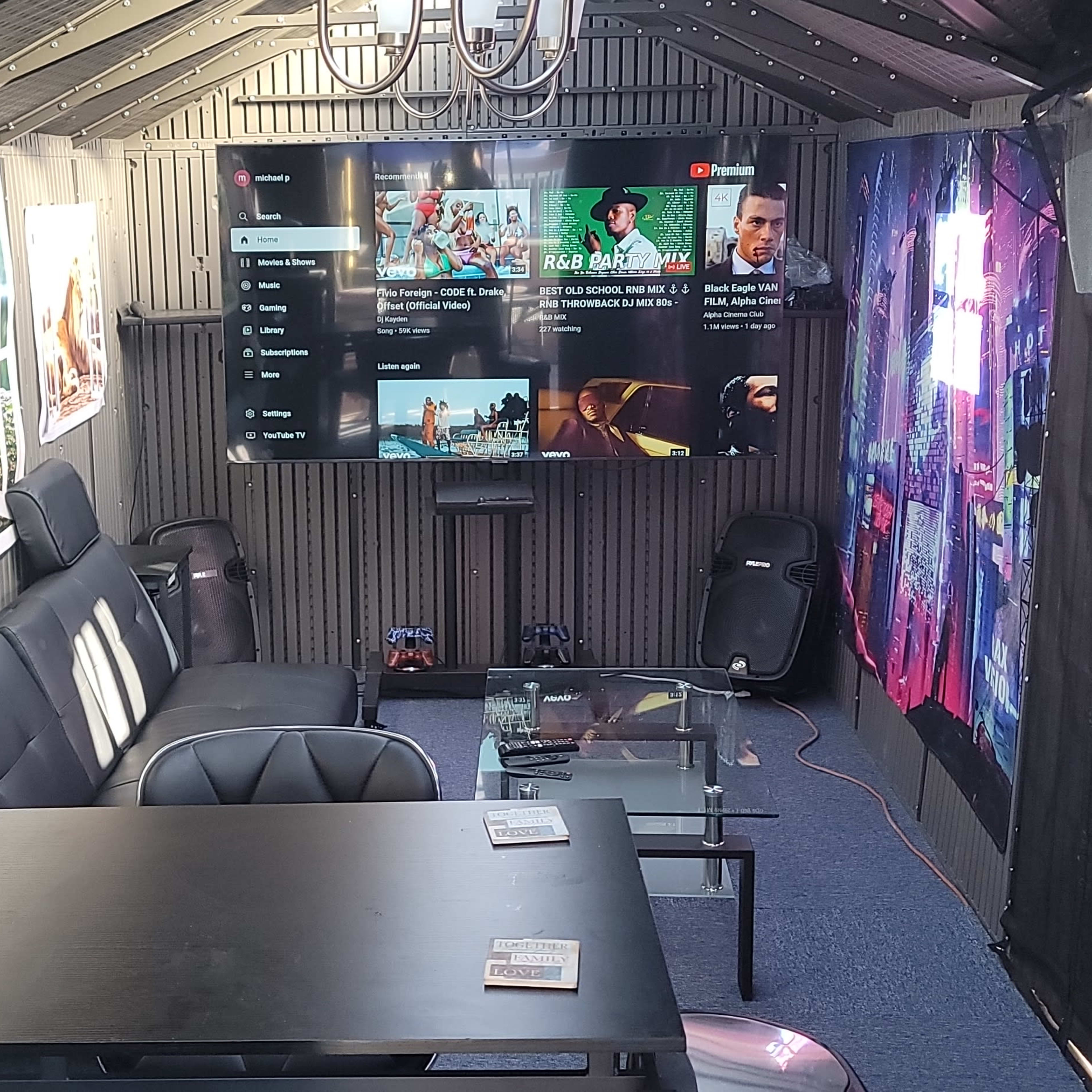 The interior of a shed-style room features a seating area with a black sofa, a large TV mounted on the wall, and decorative wall panels.