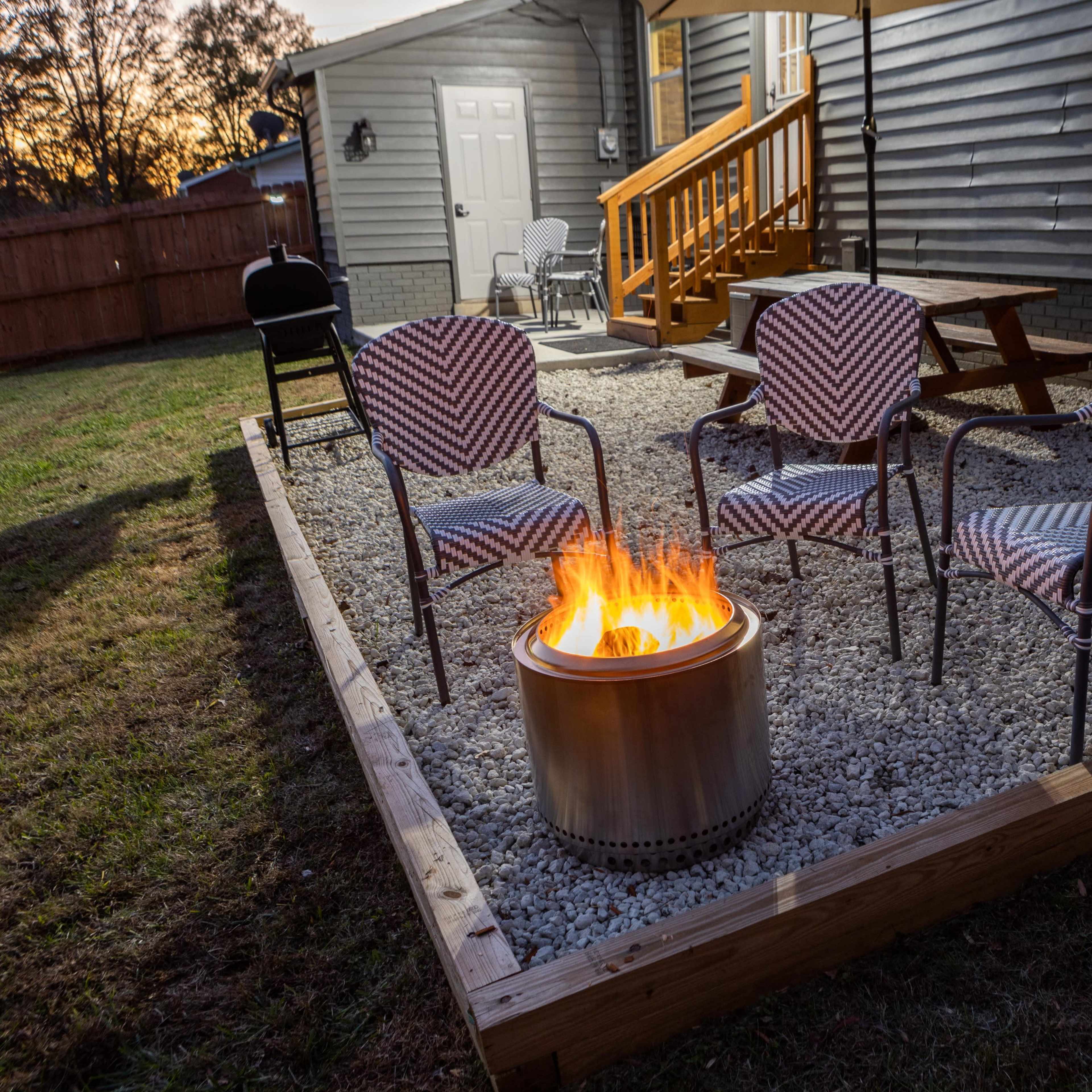 A fire pit surrounded by four chairs is set up on a gravel patio next to a house at dusk.