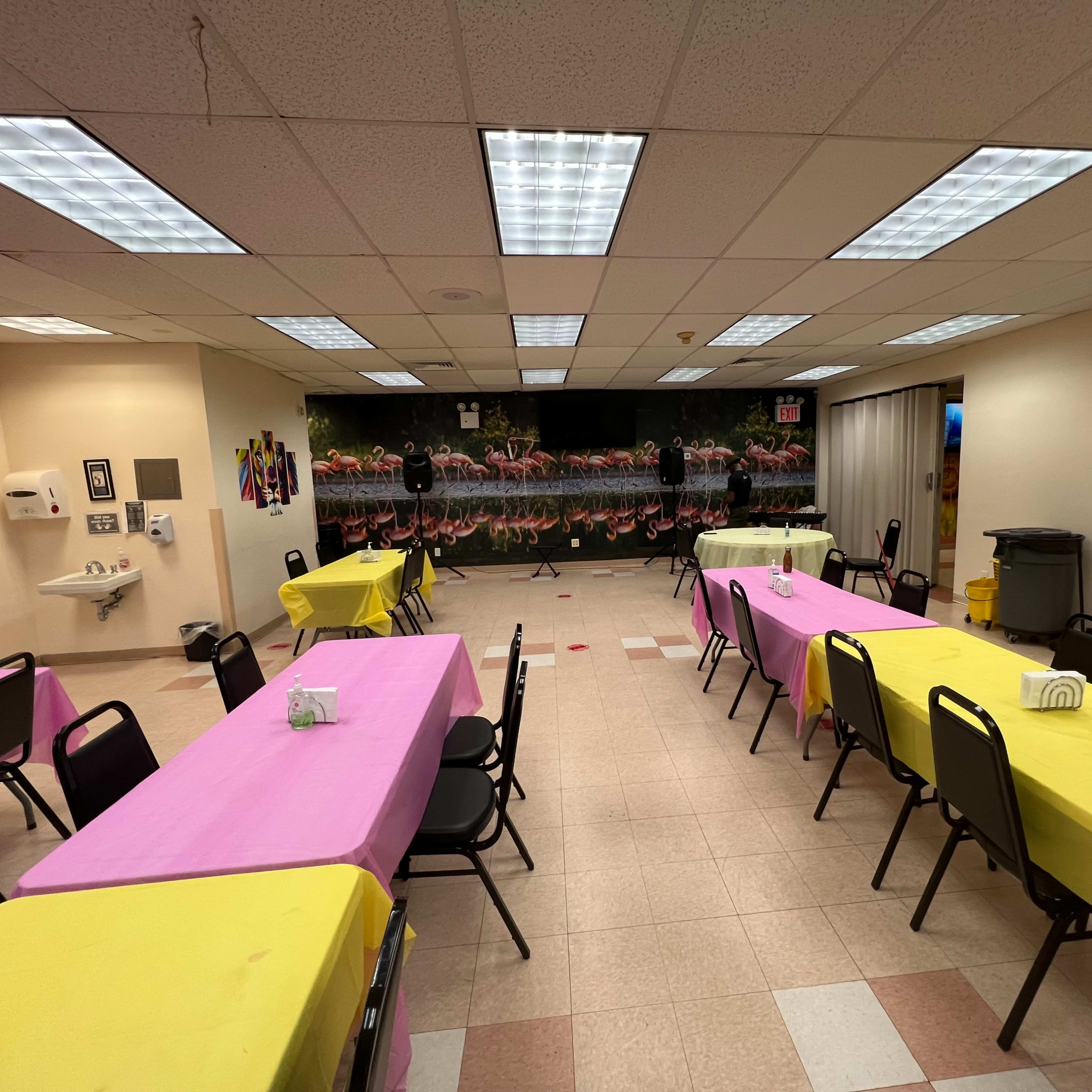 A large room set up for an event, featuring tables covered in pink and yellow tablecloths with a decorative mural on the back wall.