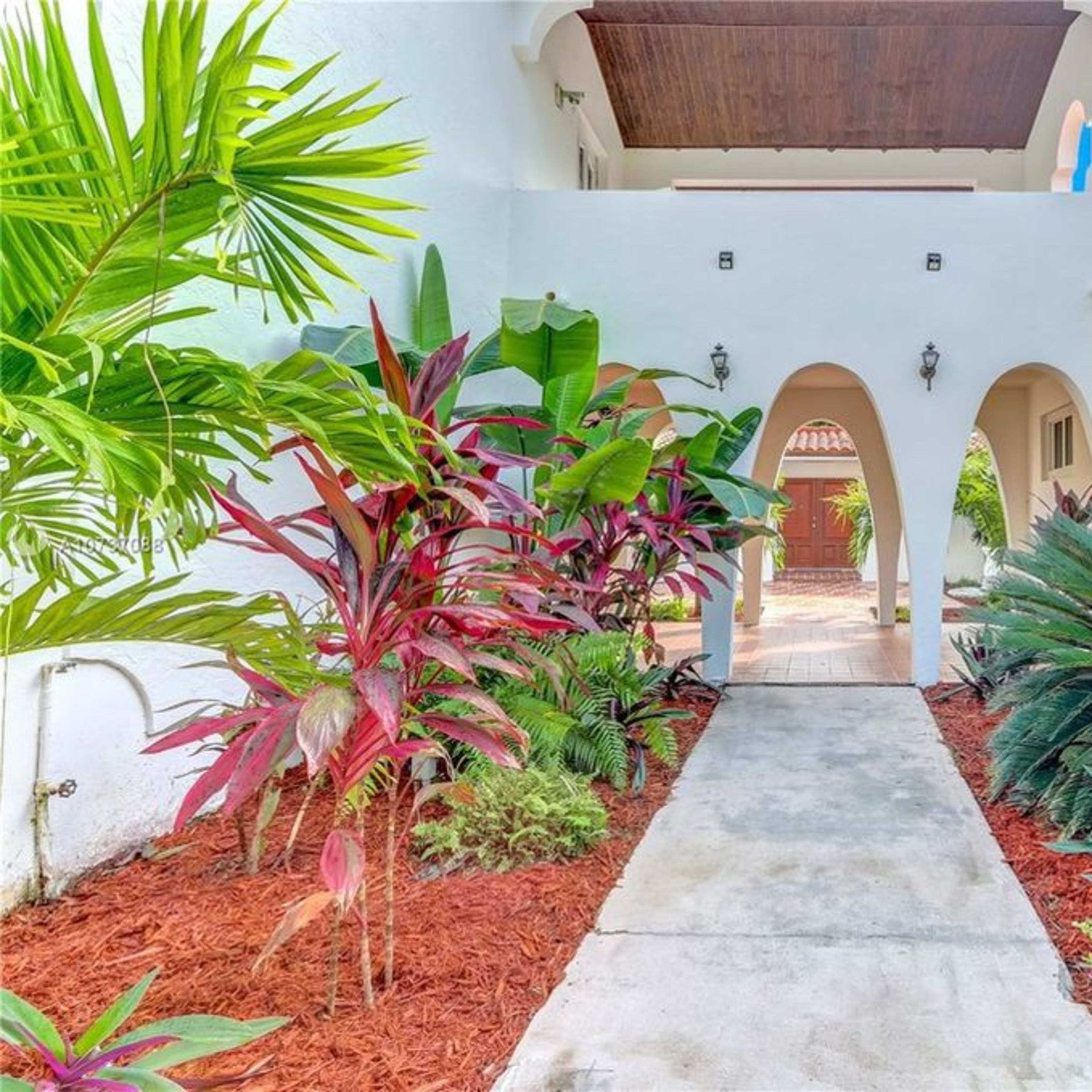 A pathway leads through a landscaped entrance with tropical plants and an arched entryway to a courtyard.