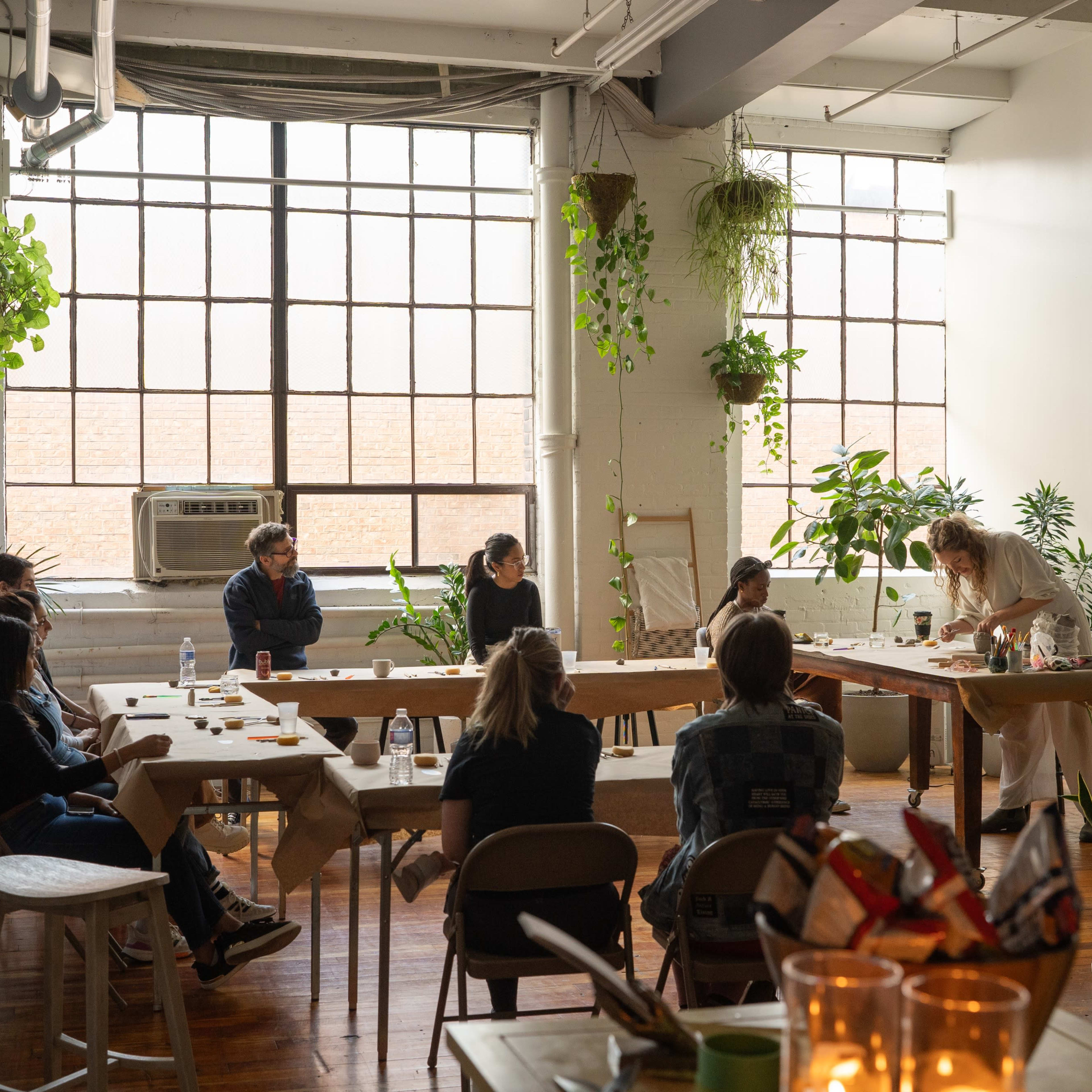 A diverse group of people gathers around a long table in a brightly lit, plant-filled room for a collaborative activity.