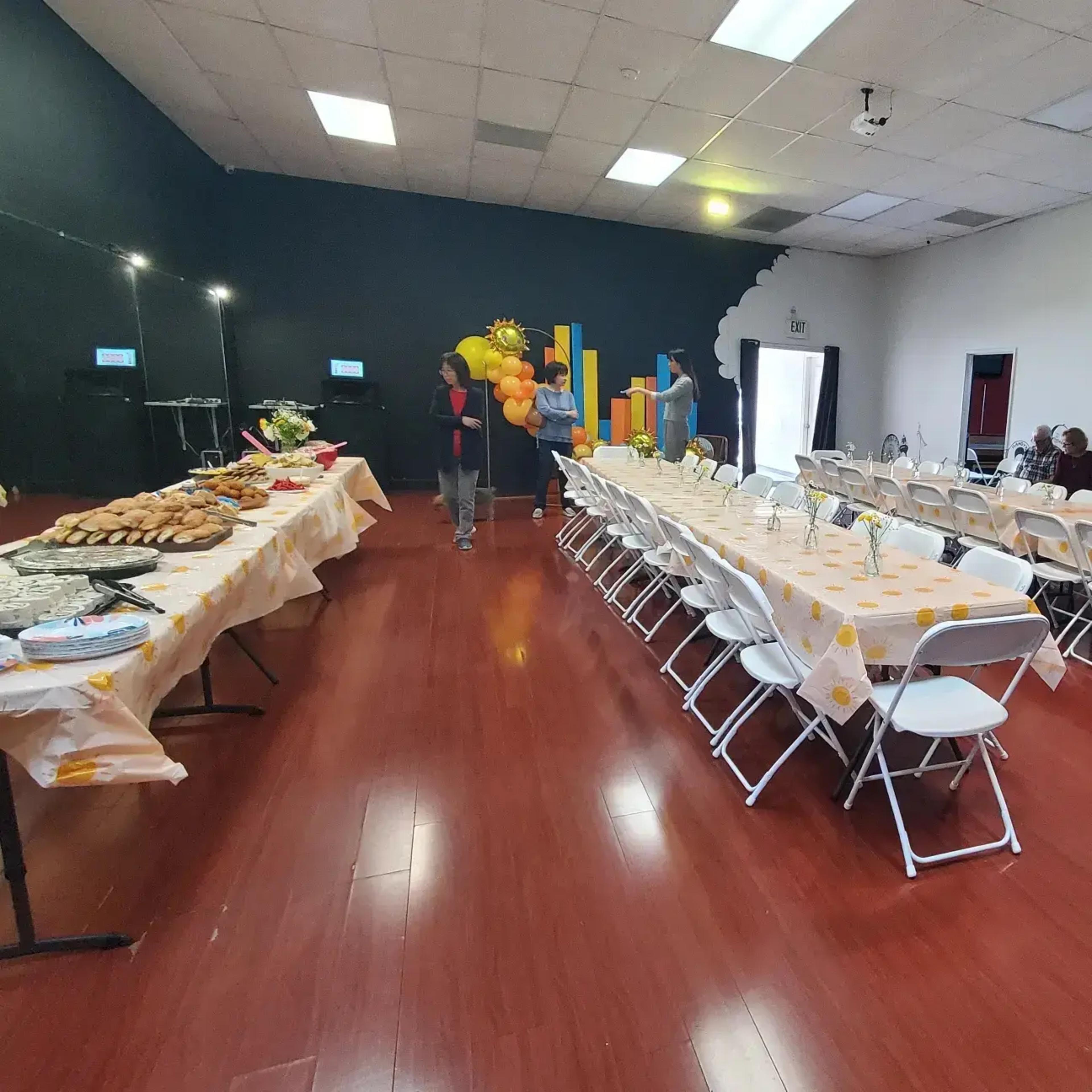 A banquet room features two long tables covered with food and decorations, while balloons and a backdrop are arranged against a dark wall.