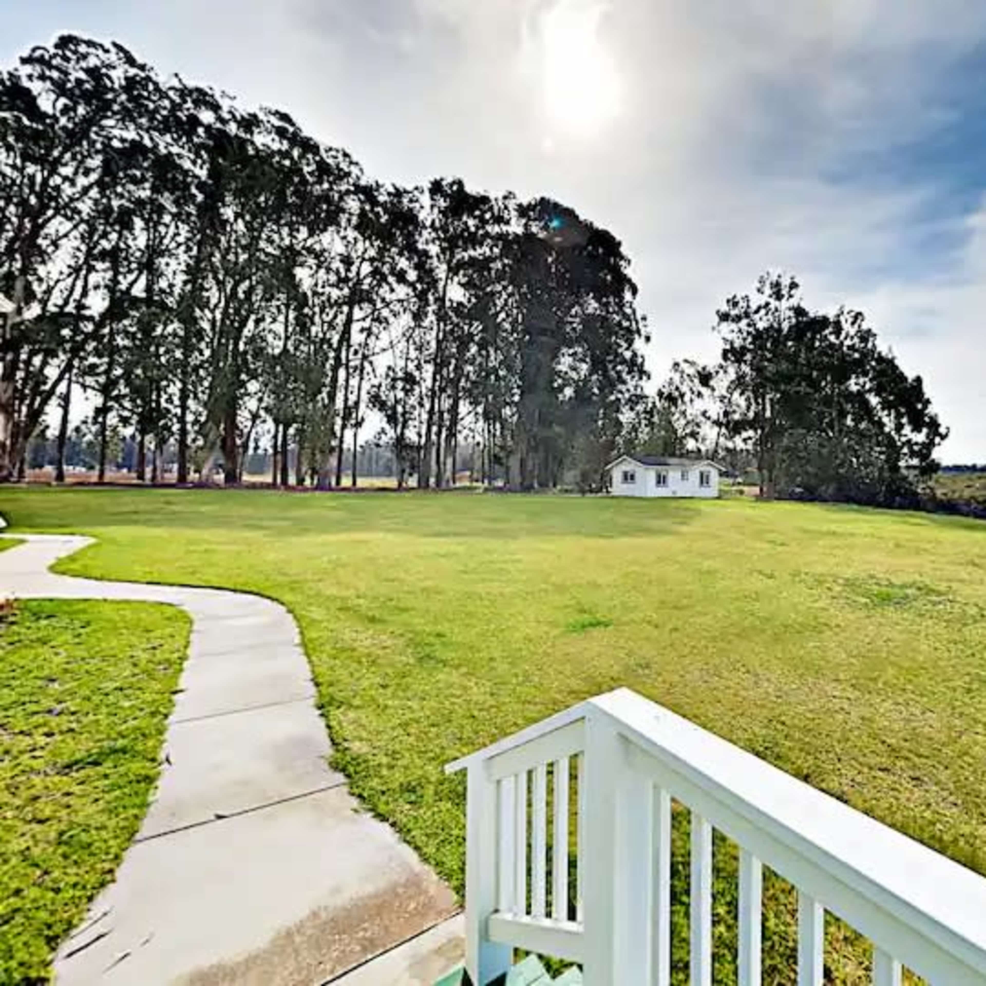 A pathway leads to a white house surrounded by tall trees and open grassy fields under a bright sky.