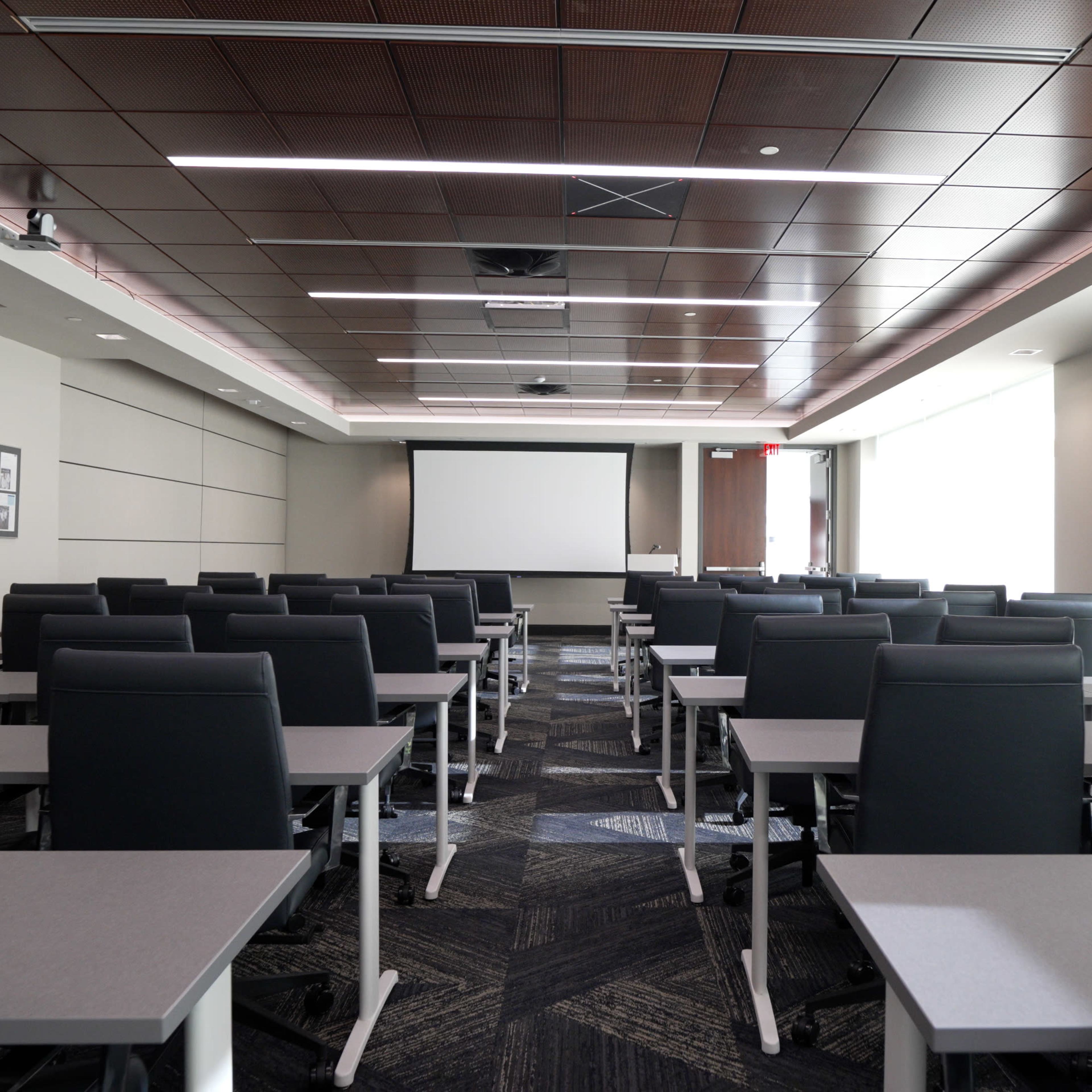 A spacious meeting room is set up with rows of gray tables and black chairs facing a large projection screen.