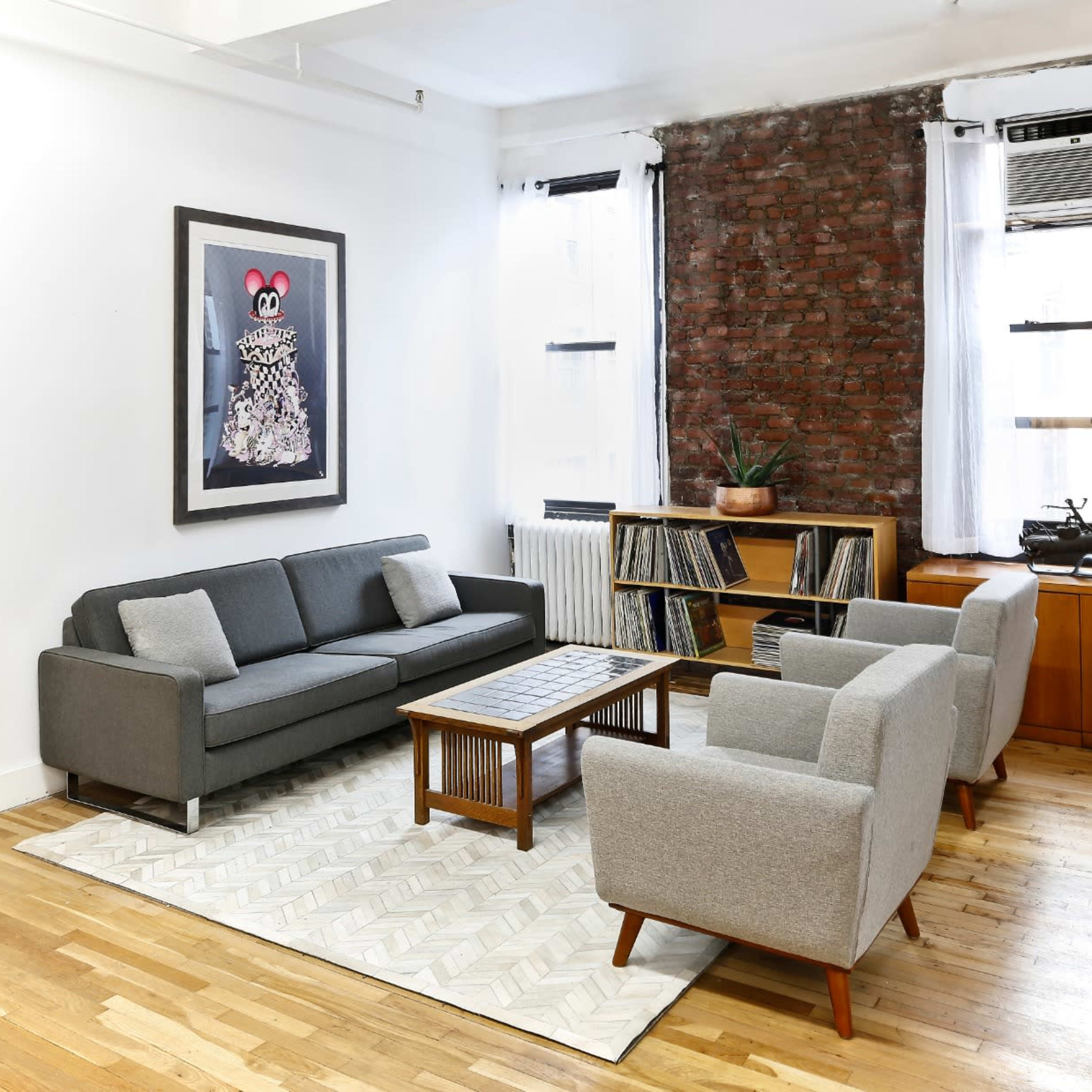 A modern living room featuring a gray sectional sofa, two gray armchairs, a wooden coffee table, a bookshelf with vinyl records, and a large framed artwork on the wall against a brick backdrop.