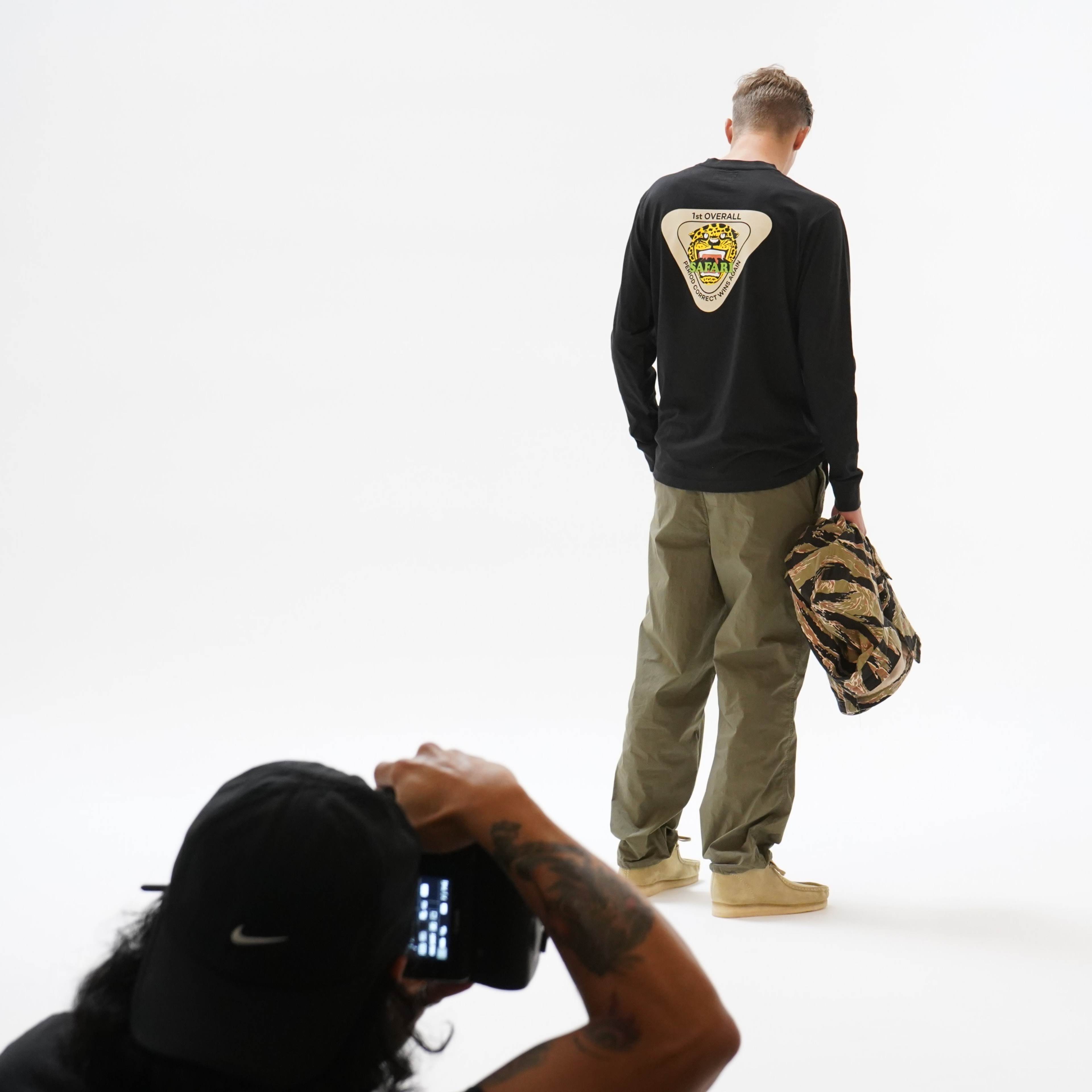 A person wearing a black long-sleeve shirt and olive green pants stands with their back to the camera while holding a camouflaged bag, as another individual takes a photograph.
