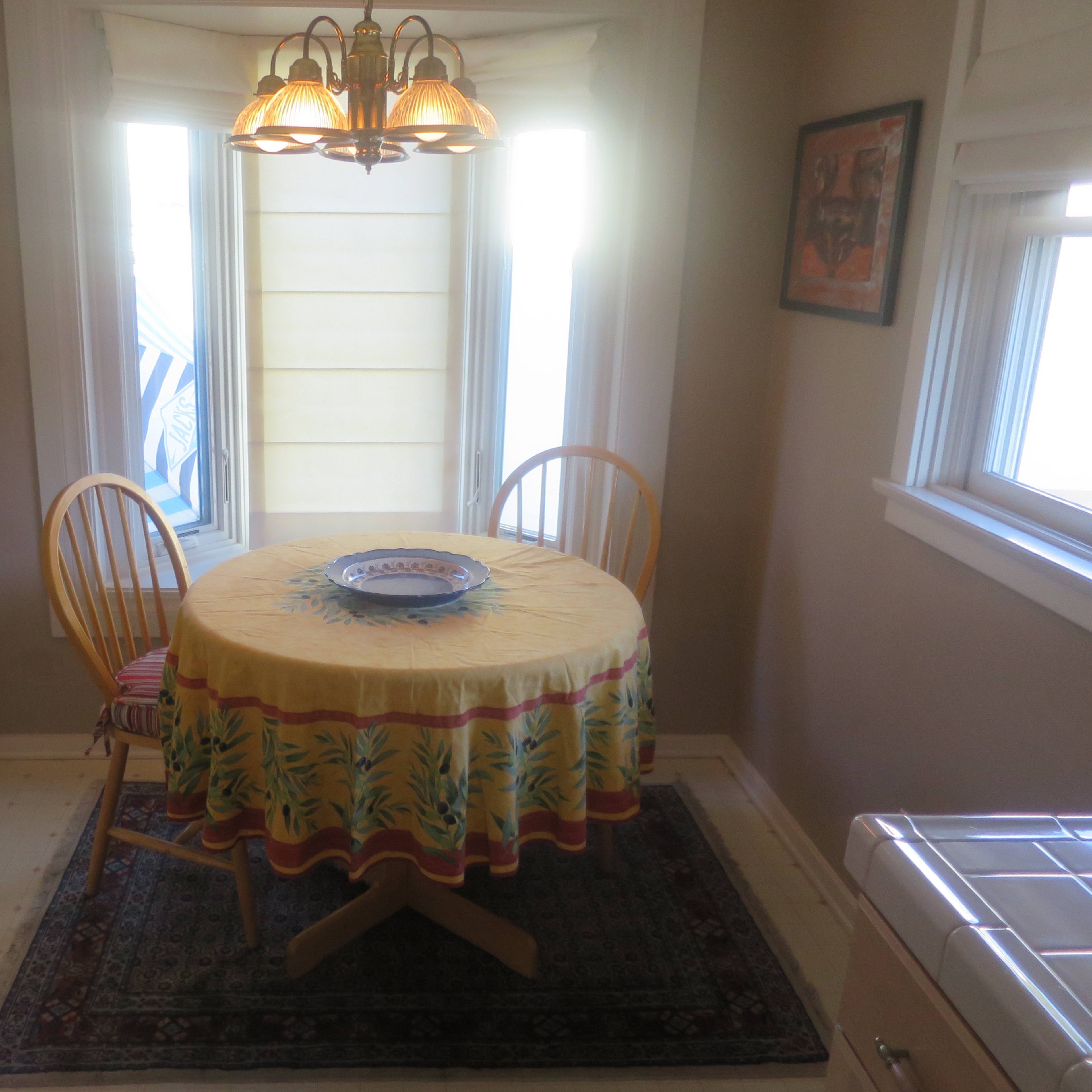 A small dining area features a round table with a yellow patterned tablecloth and two wooden chairs, surrounded by windows allowing natural light.