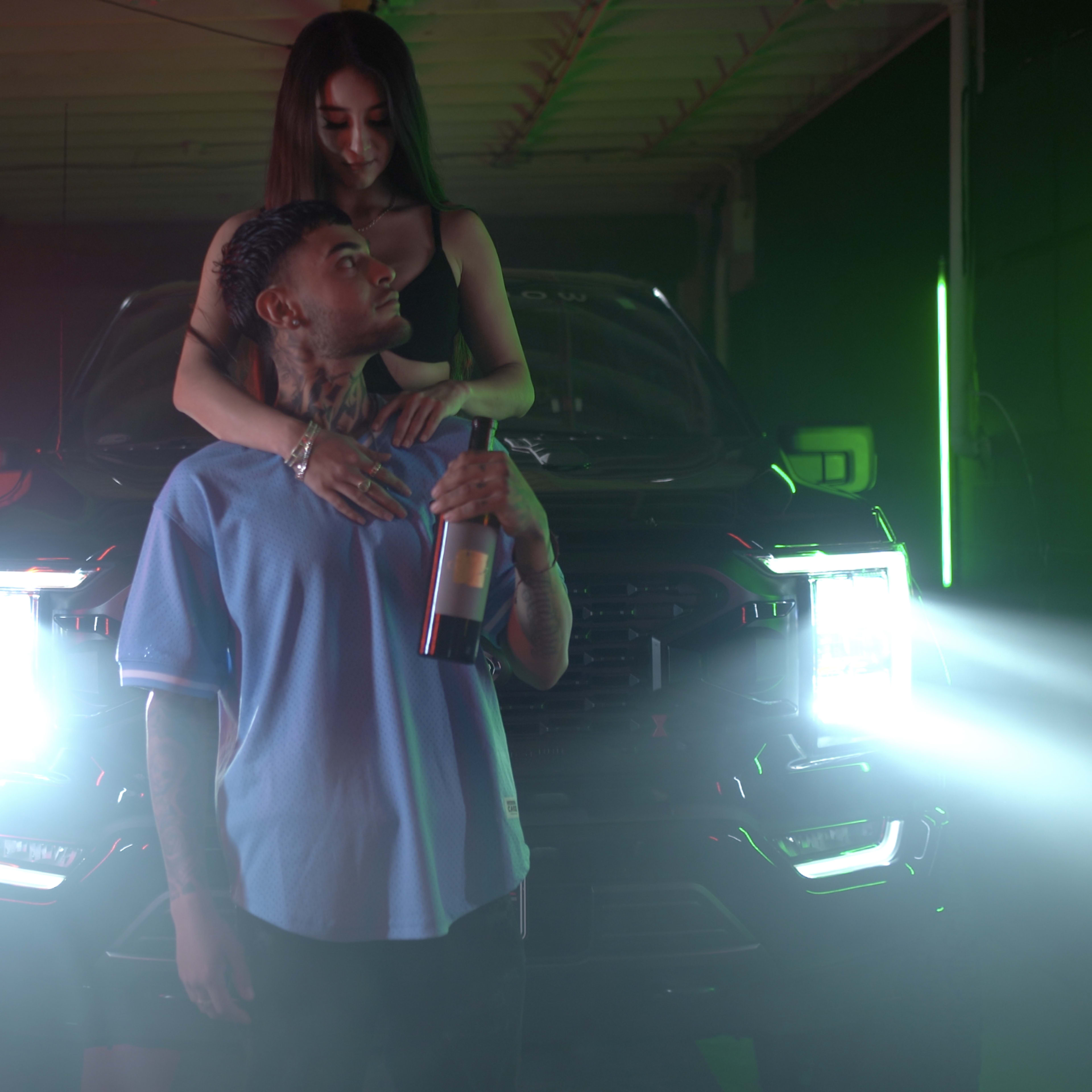A man holds a bottle while a woman embraces him in front of a car with bright headlights in a dimly lit garage.