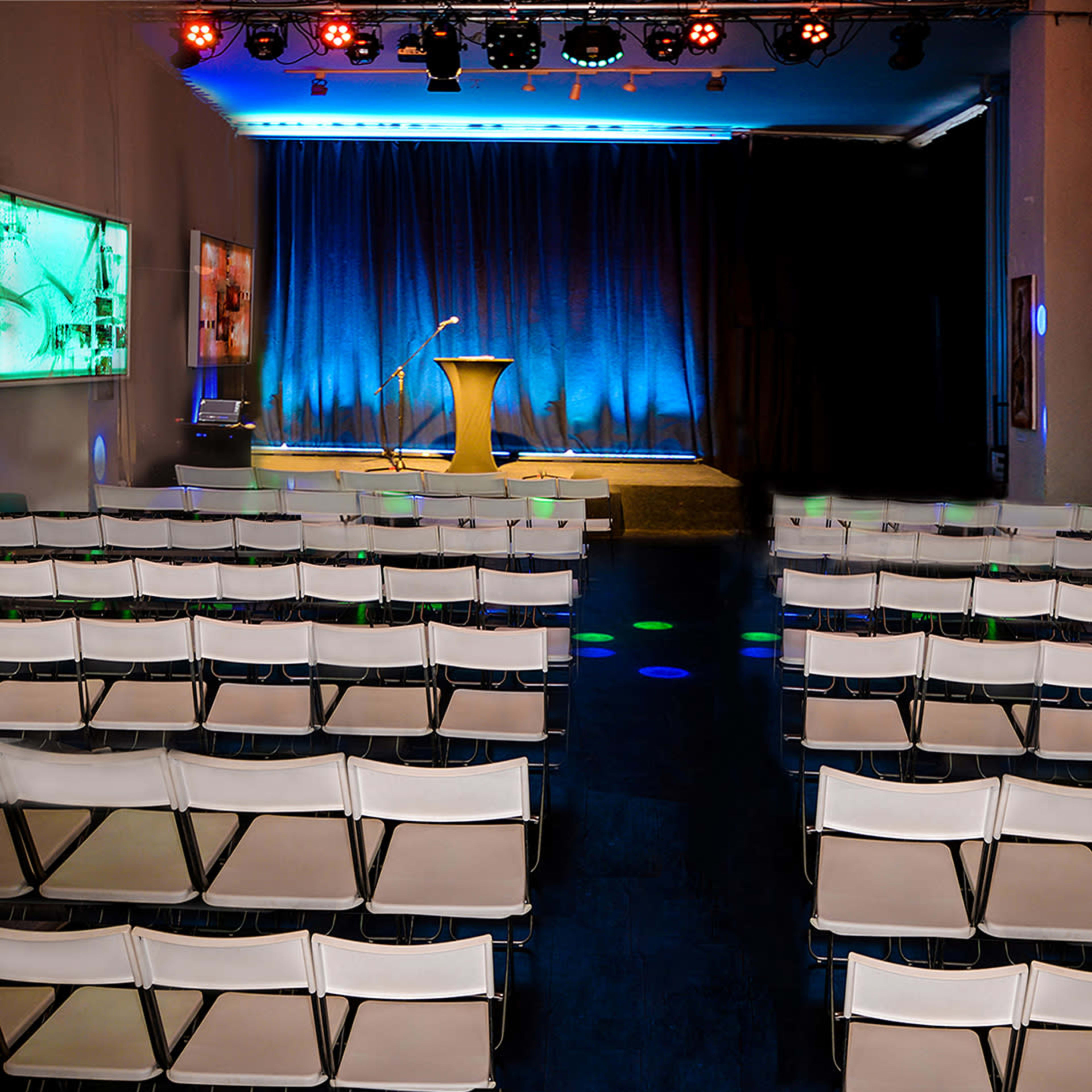 A stage with a podium and microphone is set at the front of a room filled with rows of empty folding chairs under blue and colorful lights.