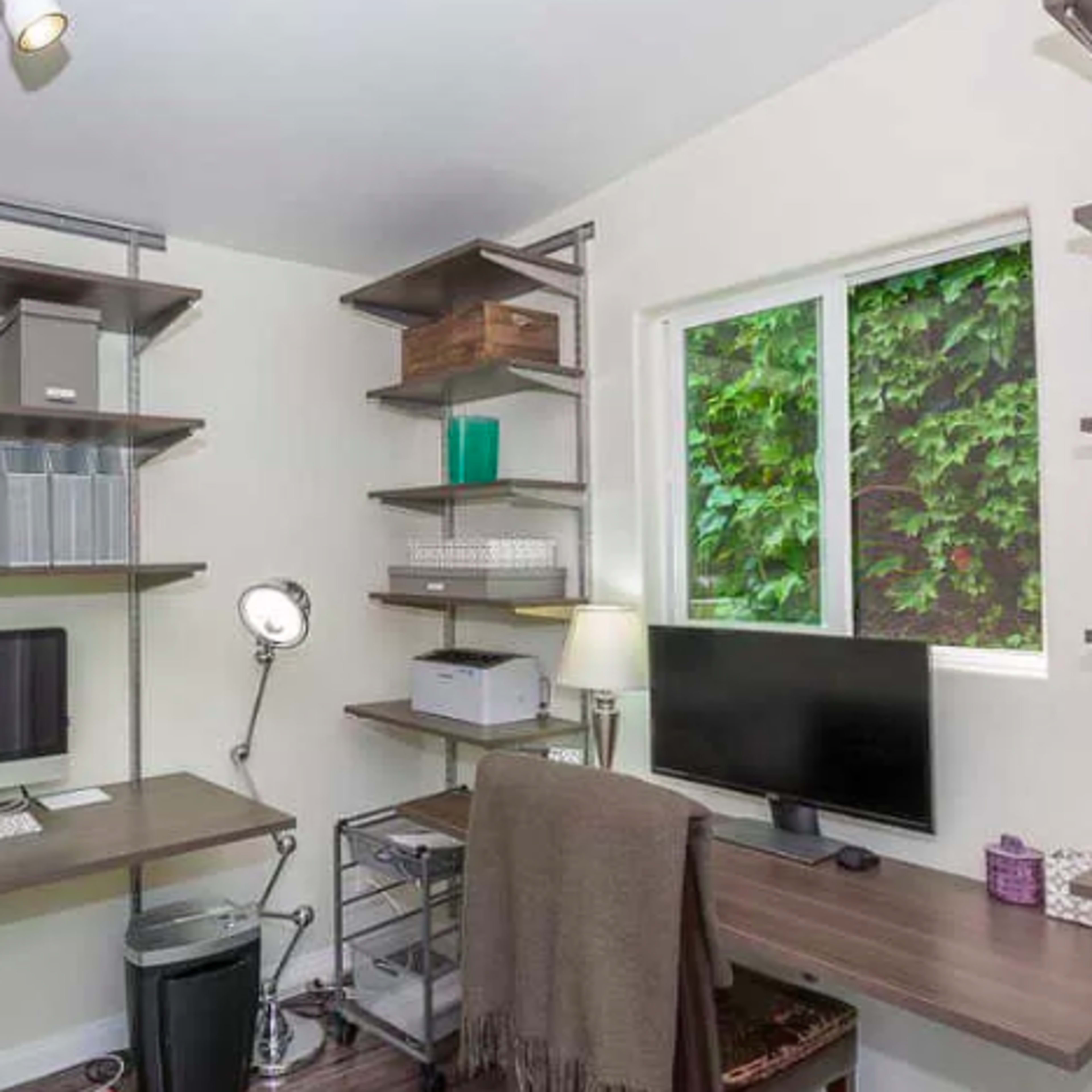 A workspace features two desks with computers, organized shelves on the walls, and a window overlooking greenery.