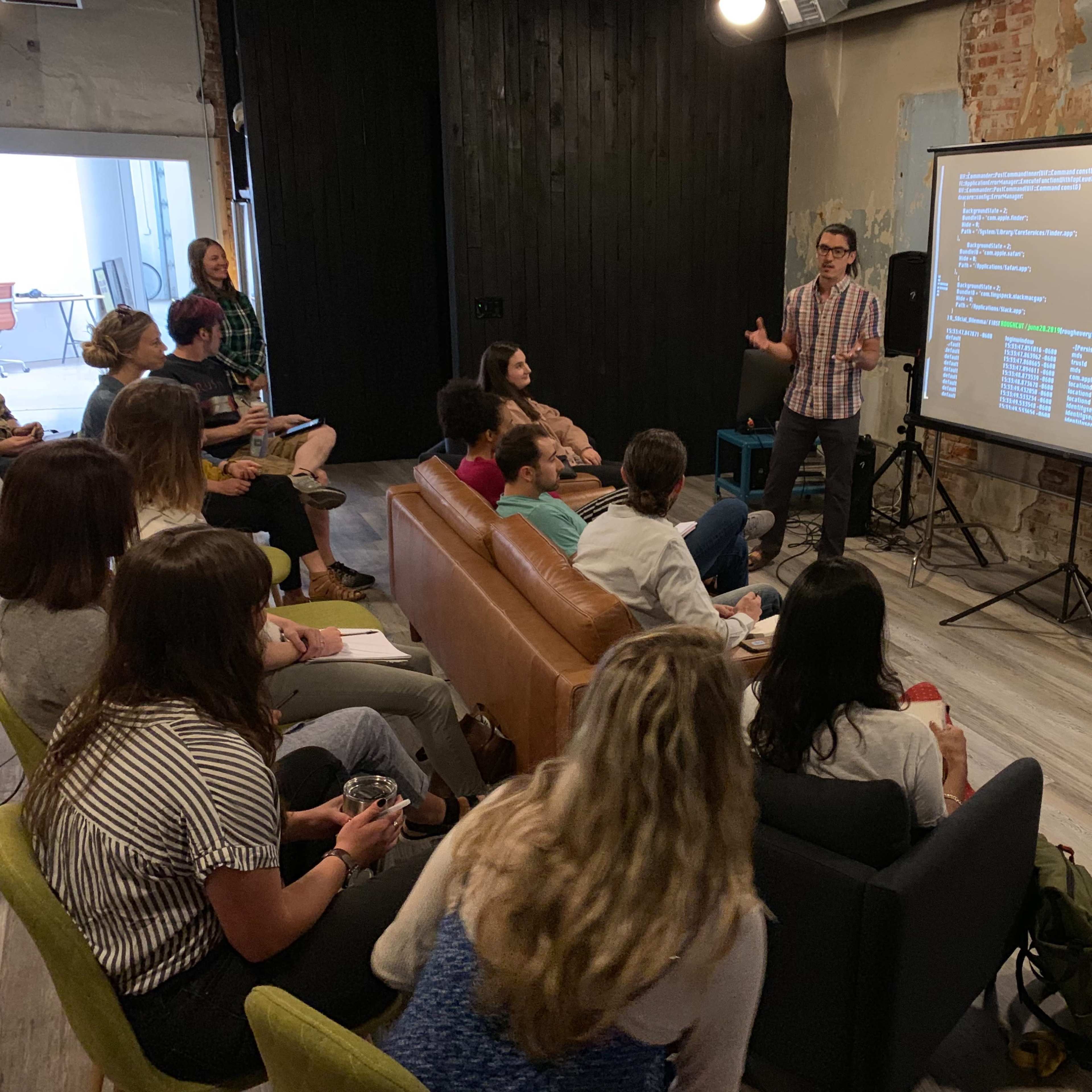 A presenter stands in front of an audience in a creative workspace, discussing information displayed on a large screen.