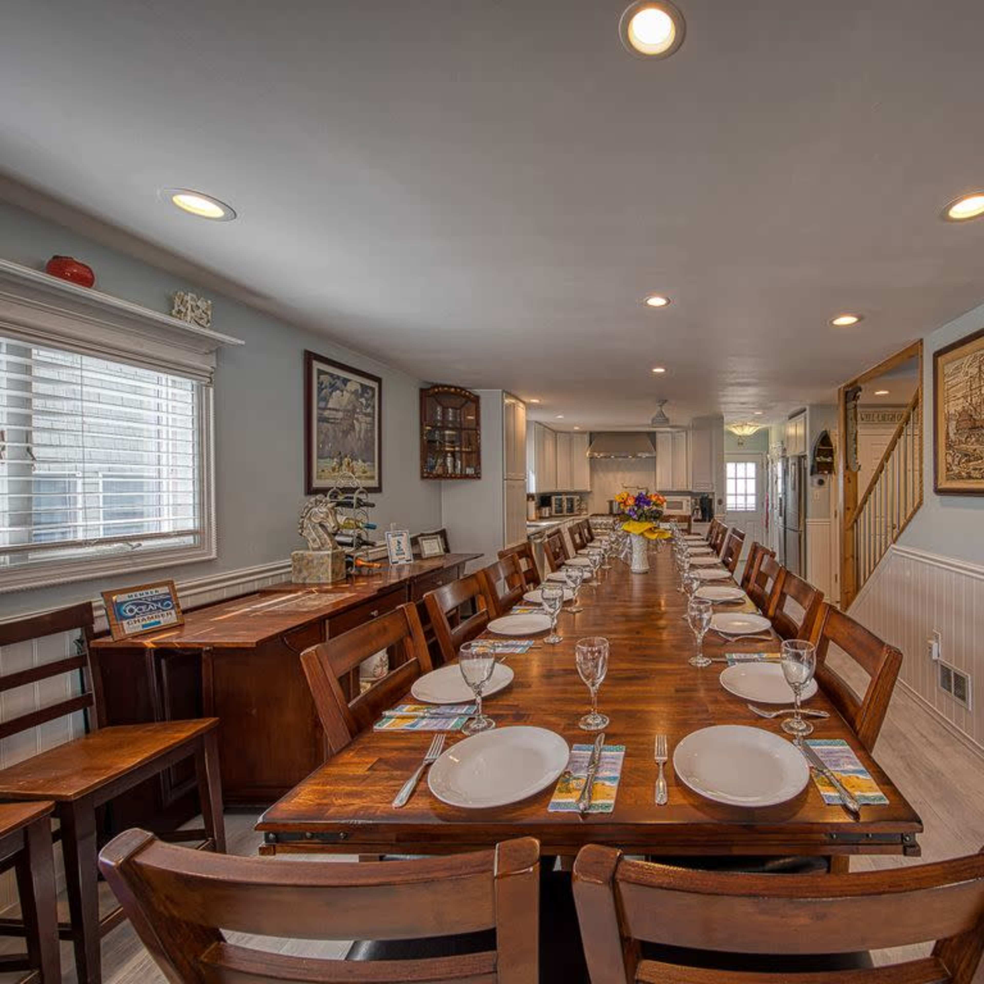 A long wooden dining table is set with plates and glasses in a well-lit dining room that leads to a kitchen and staircase.