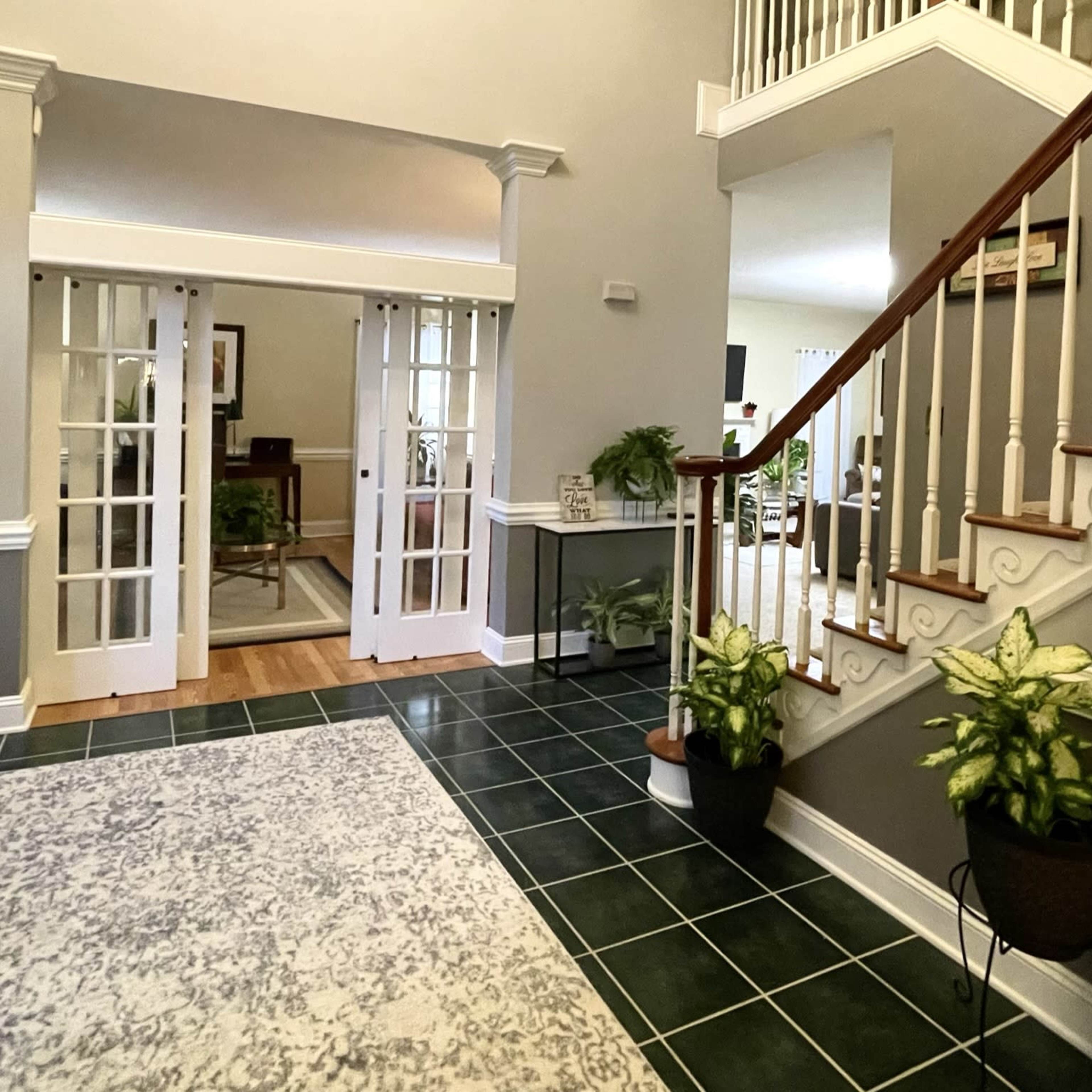 A spacious foyer with green tiled flooring, a staircase, and double doors leading to a living area, adorned with potted plants.