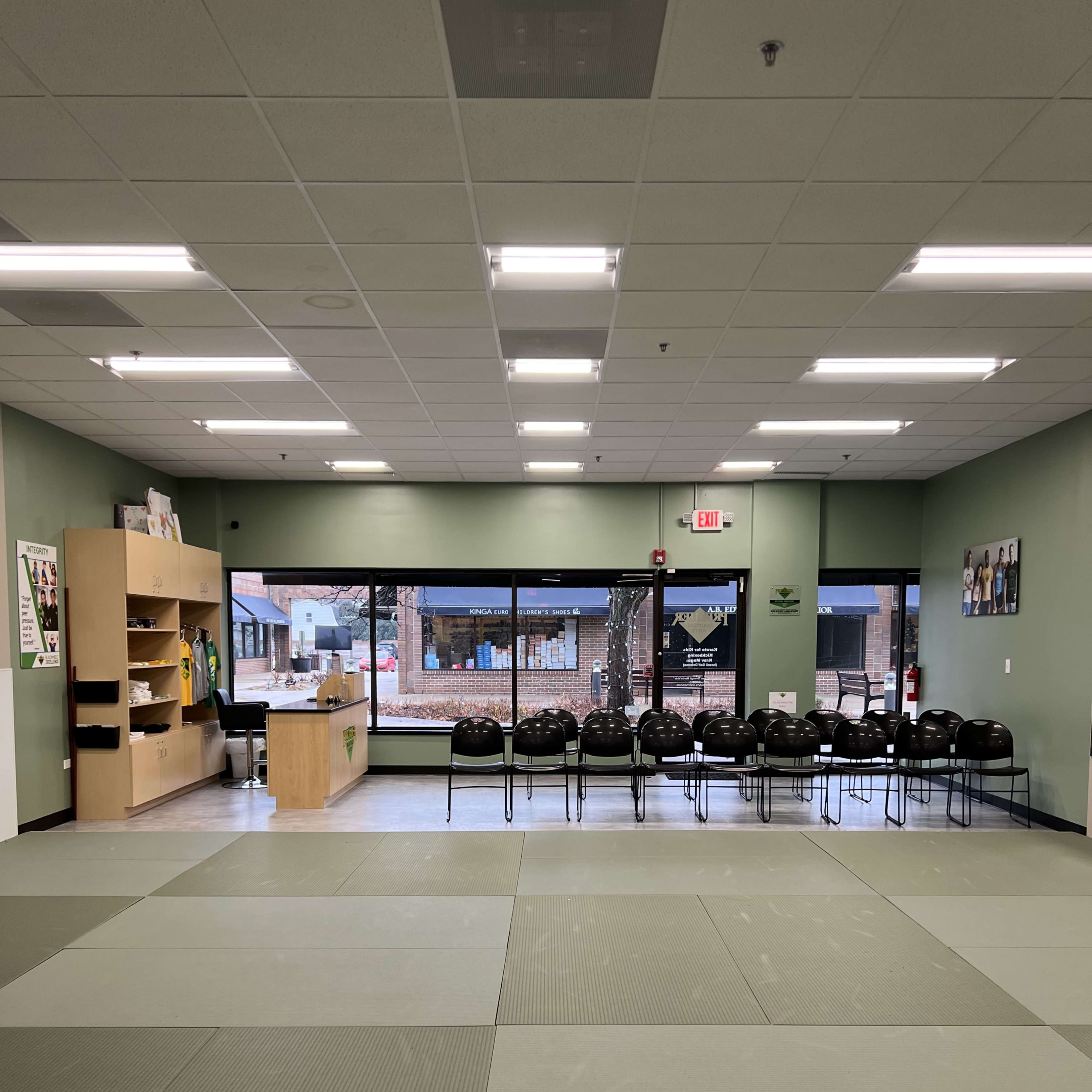 A dojo with a mat area in the foreground and a row of black chairs lined up against a wall, along with a reception area and large windows in the background.