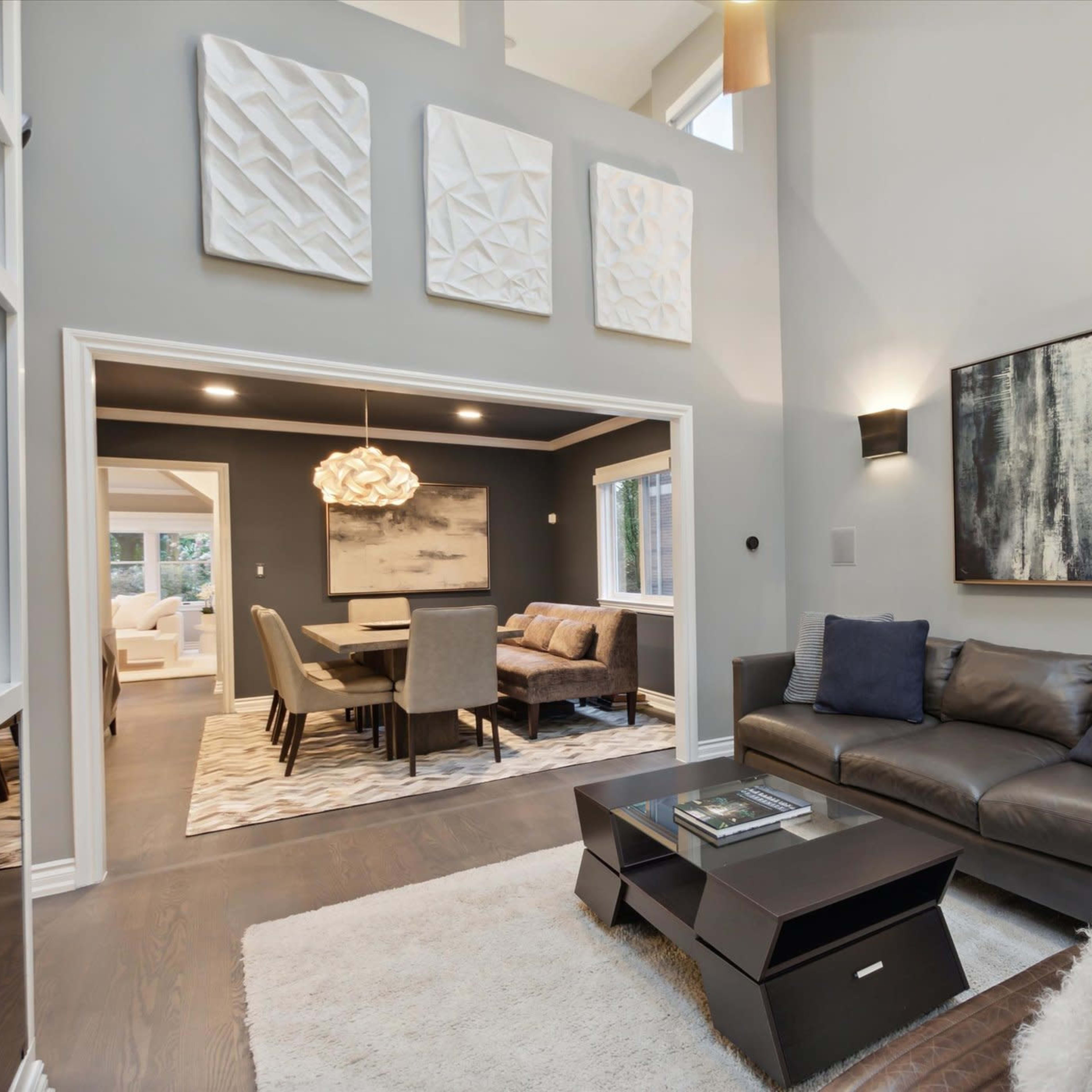 A modern living room features a dark leather sofa facing a sleek coffee table, with large artwork on the walls and a dining area visible through an archway.