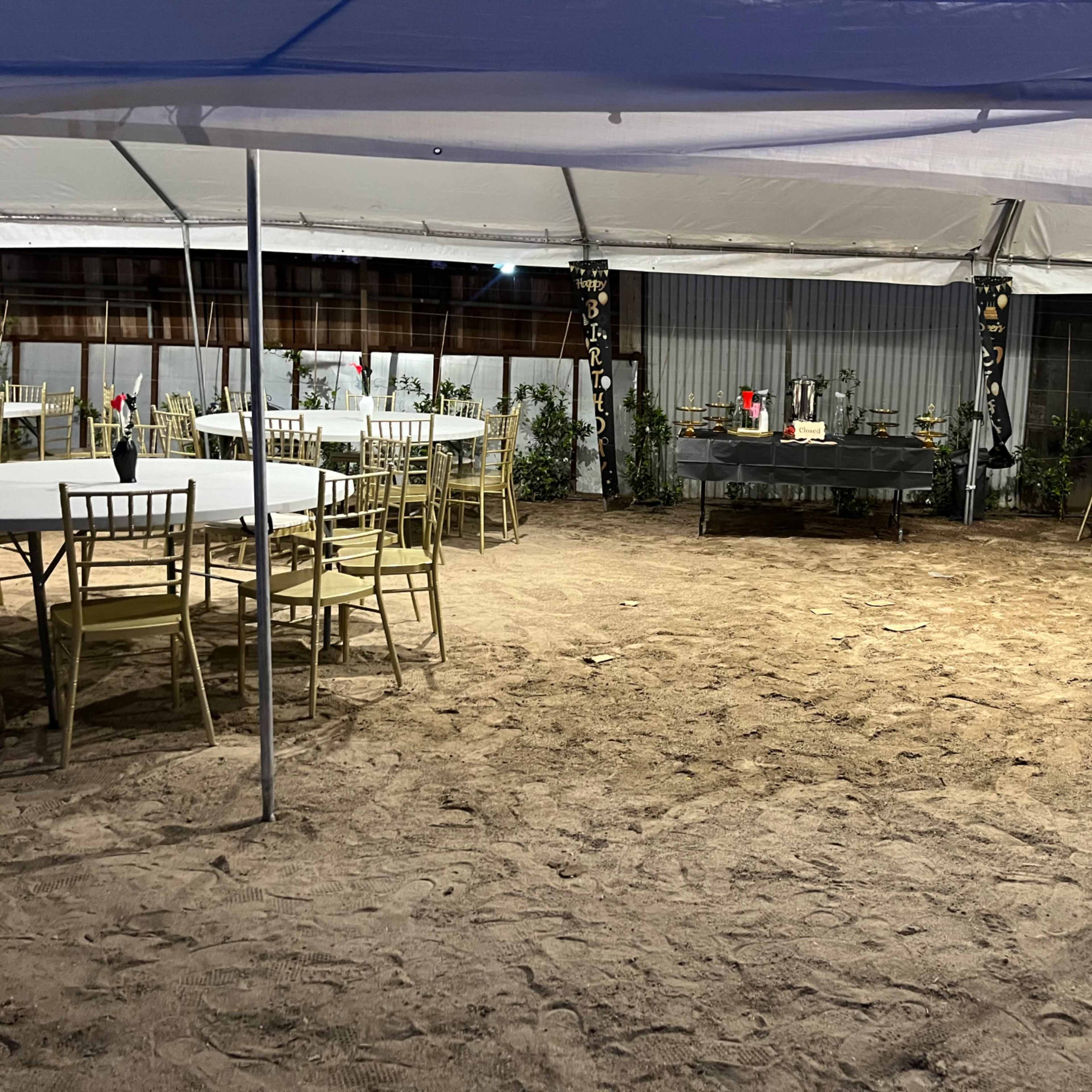 A large tented area features circular tables and chairs arranged on sandy ground, with a decorative table at the center surrounded by plants and flowers.