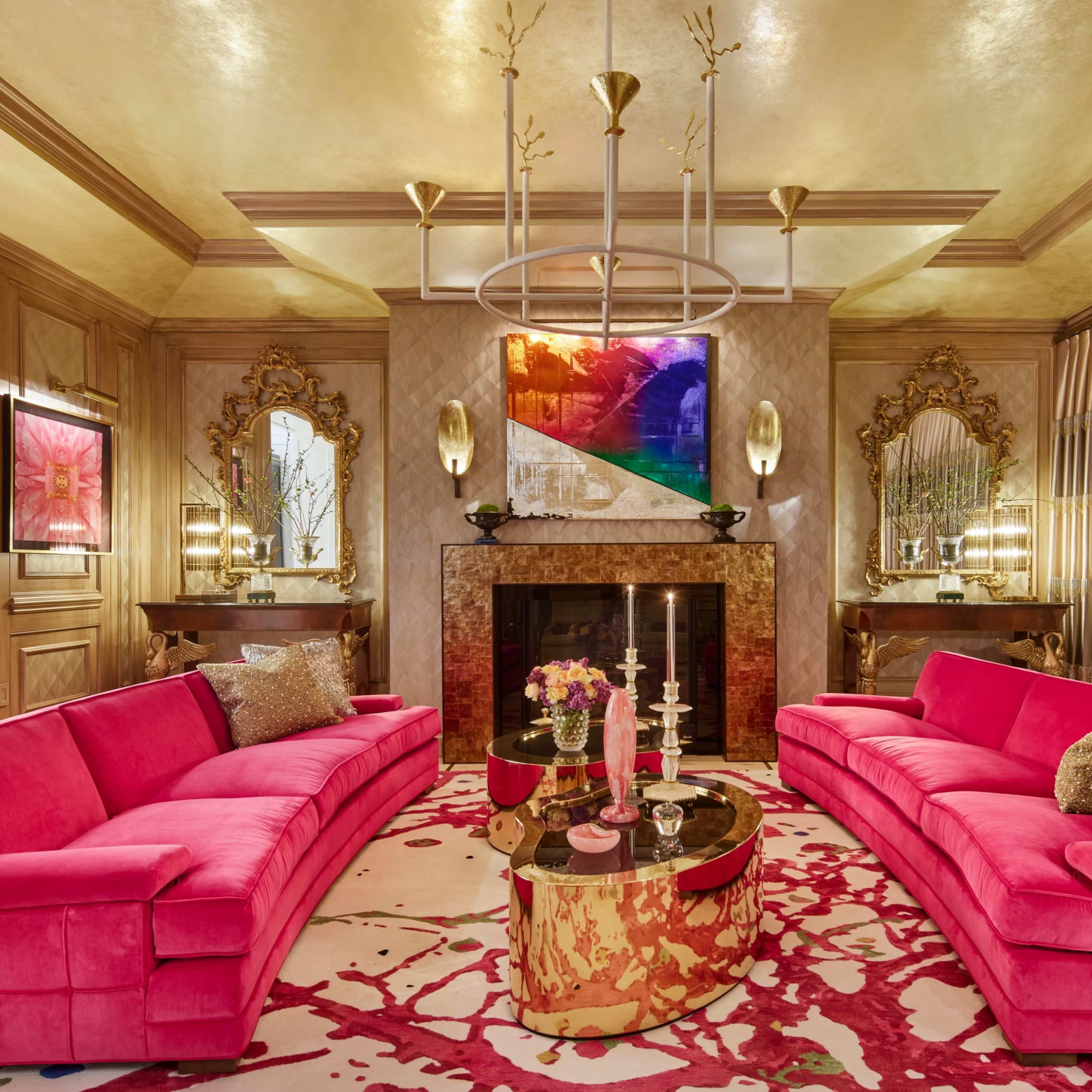 A luxurious living room features two pink sofas facing each other, a circular coffee table, a fireplace, and decorative mirrors against a gold-hued wall.