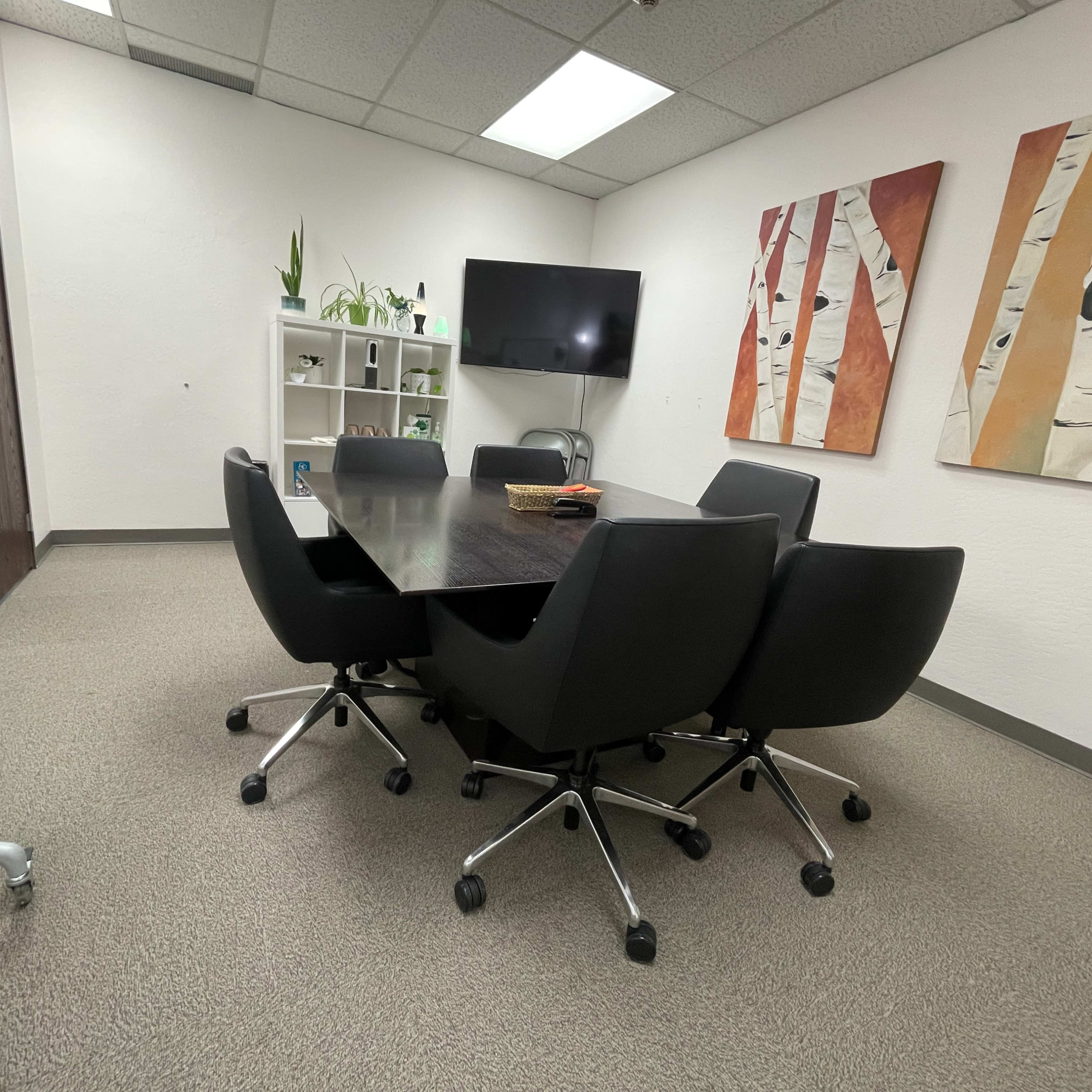 A conference room features a dark table surrounded by black chairs, with a television and shelving unit on the walls.