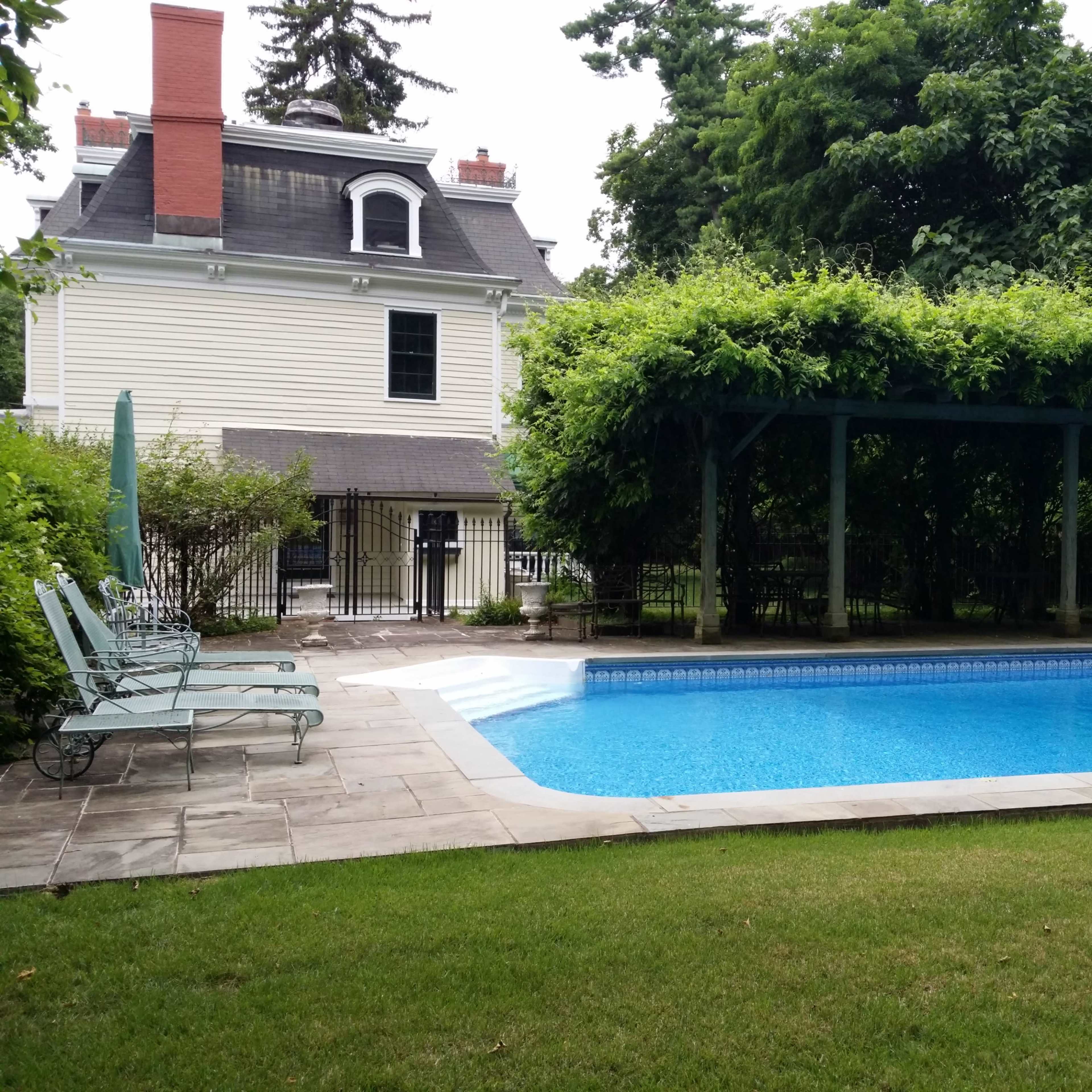 A rectangular swimming pool is surrounded by a stone patio and a grassy area, with a pergola covered in greenery nearby, and a house visible in the background.