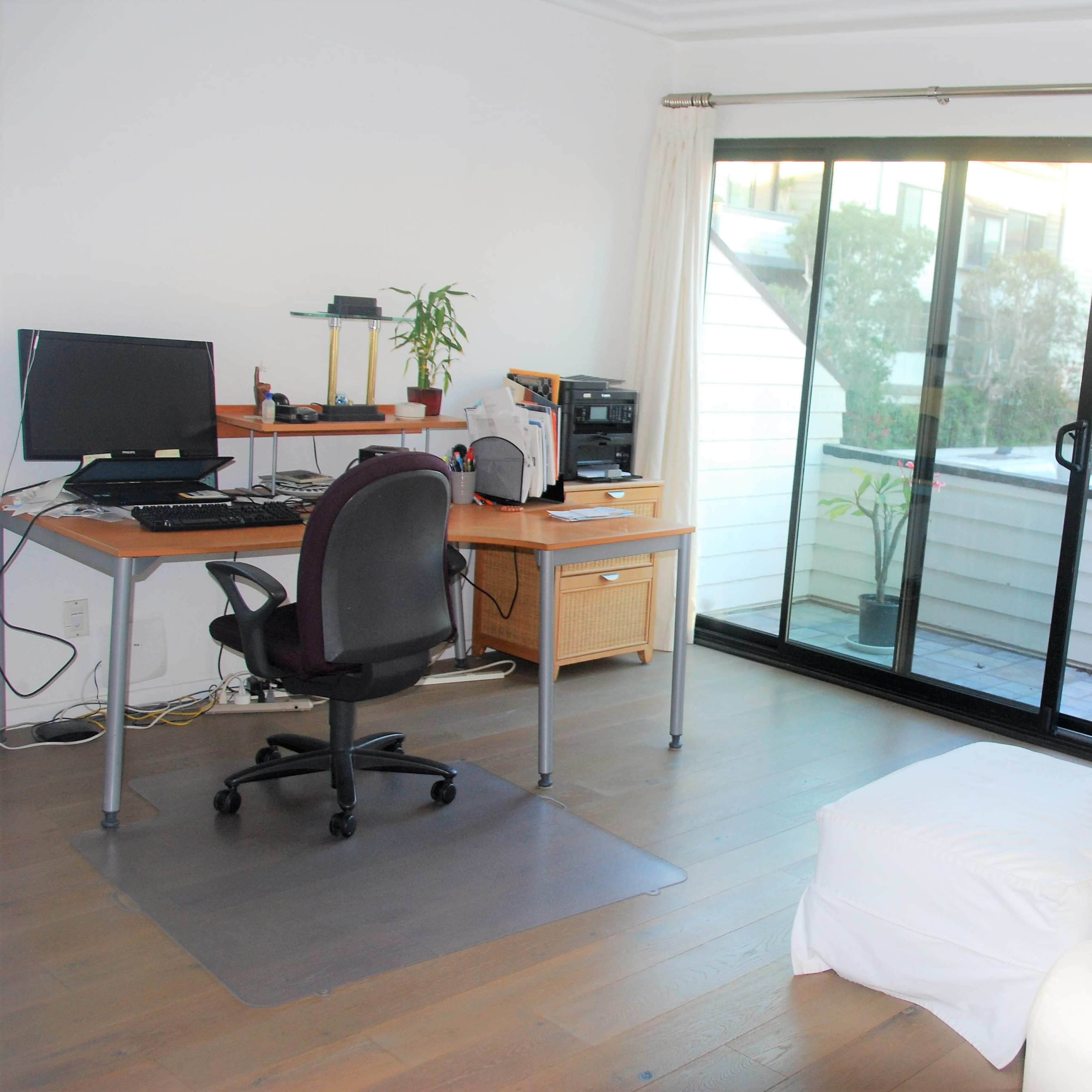 A home office with a desk, computer, and a chair, adjacent to a glass sliding door that leads to a balcony.