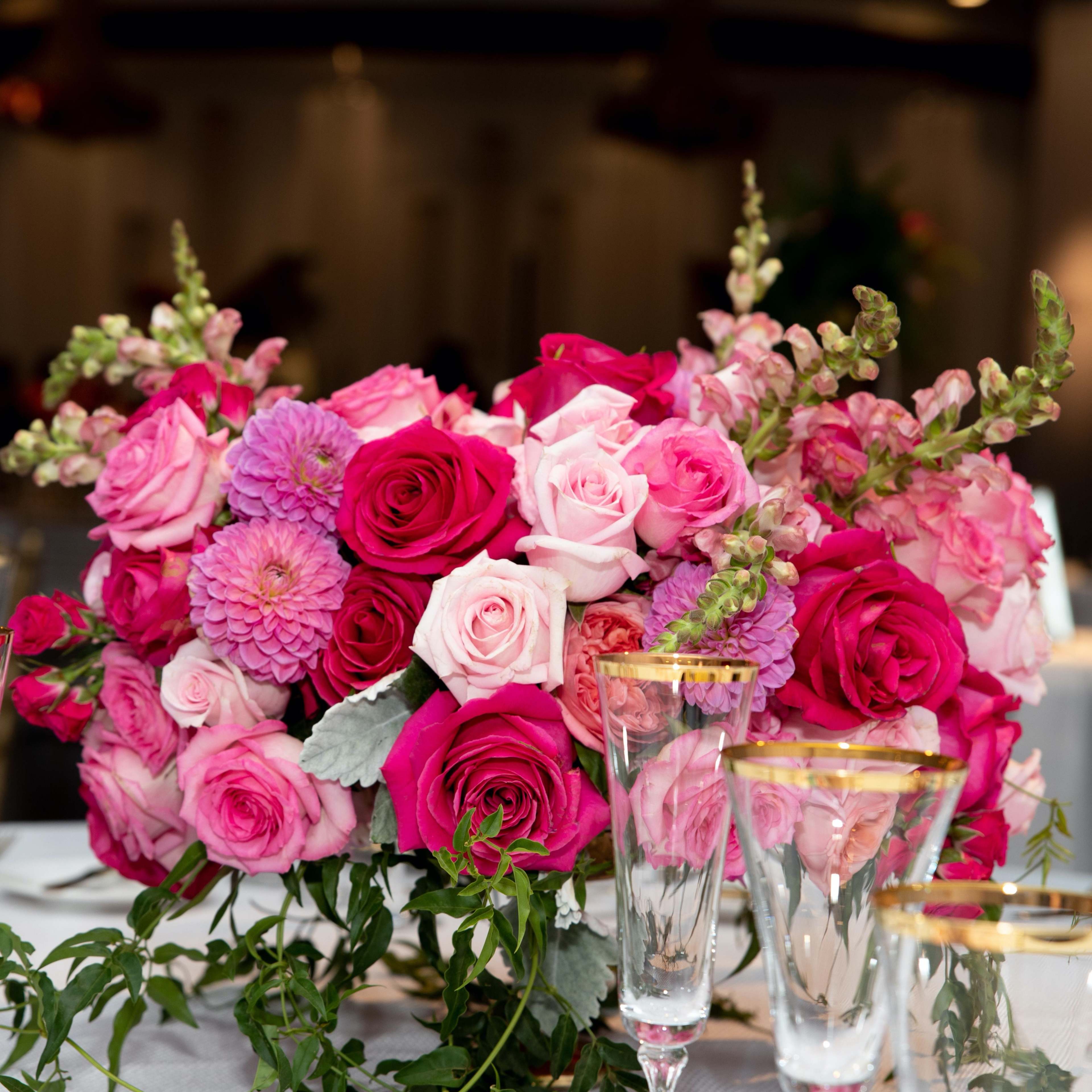A vibrant floral arrangement featuring pink and red roses, zinnias, and snapdragons is displayed at a dining table surrounded by elegant glassware.