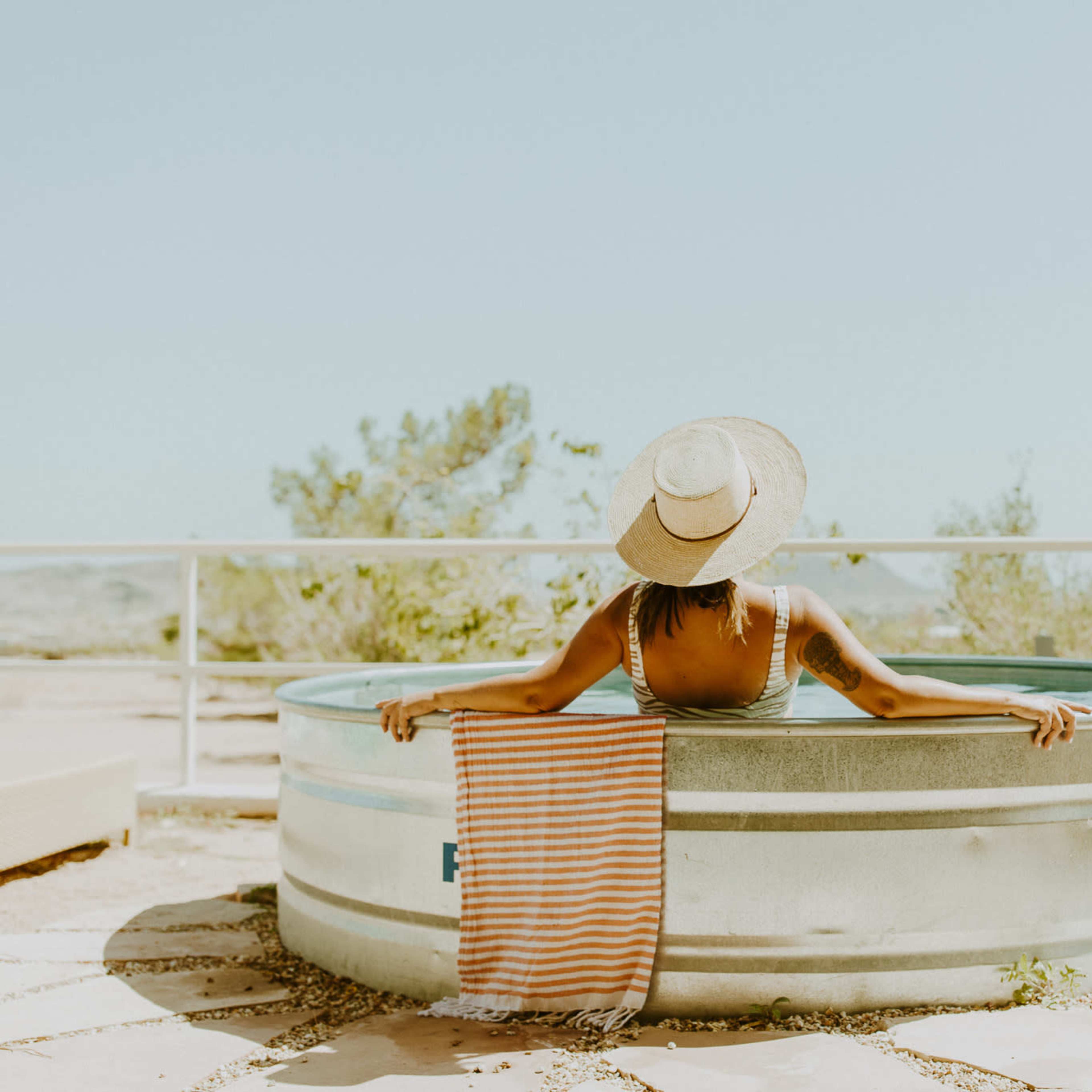A person sits in a metal bathtub outdoors, holding a striped towel, with trees and a distant landscape in the background.