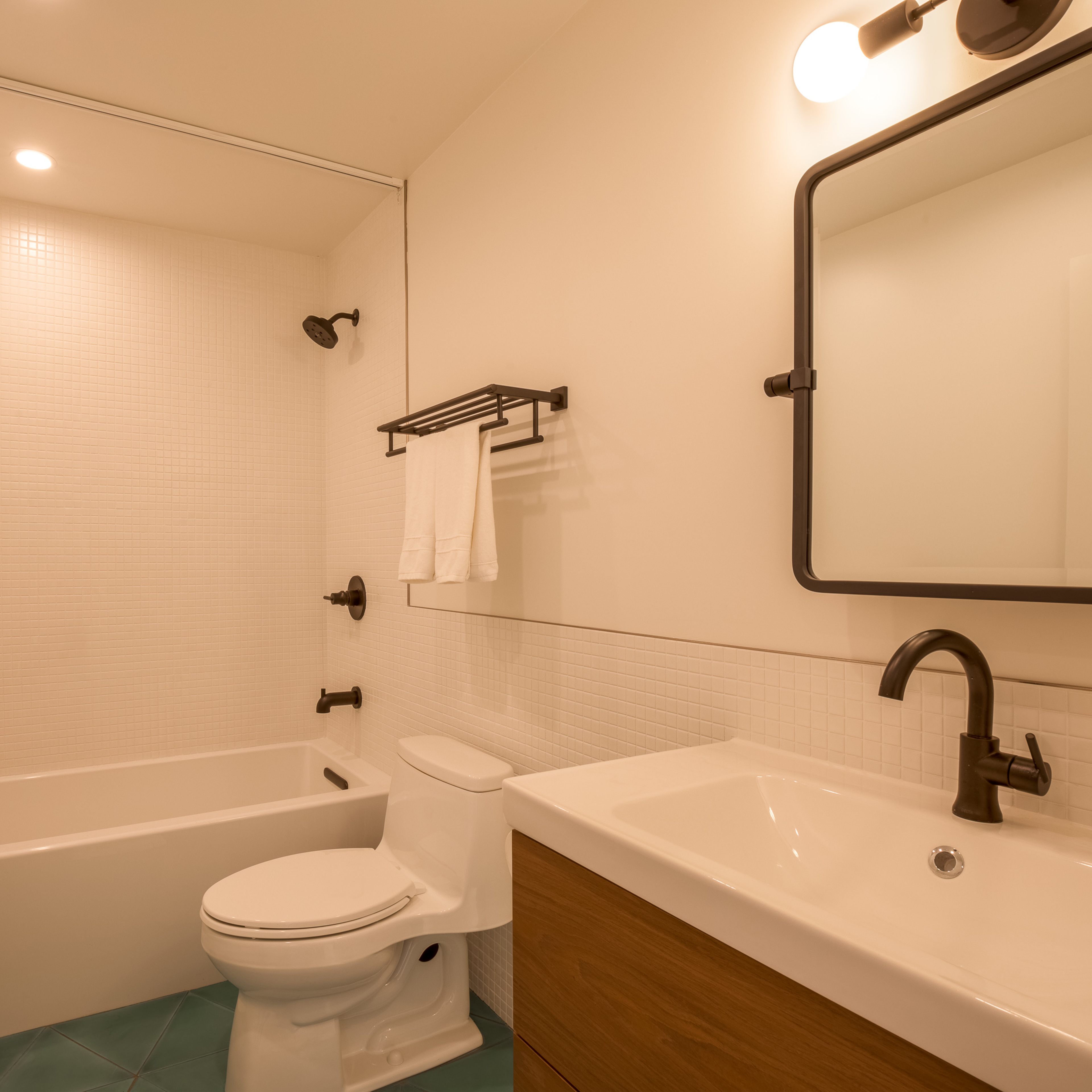 A modern bathroom featuring a bathtub with a shower, a toilet, a sink with a wooden vanity, and simple wall fixtures.