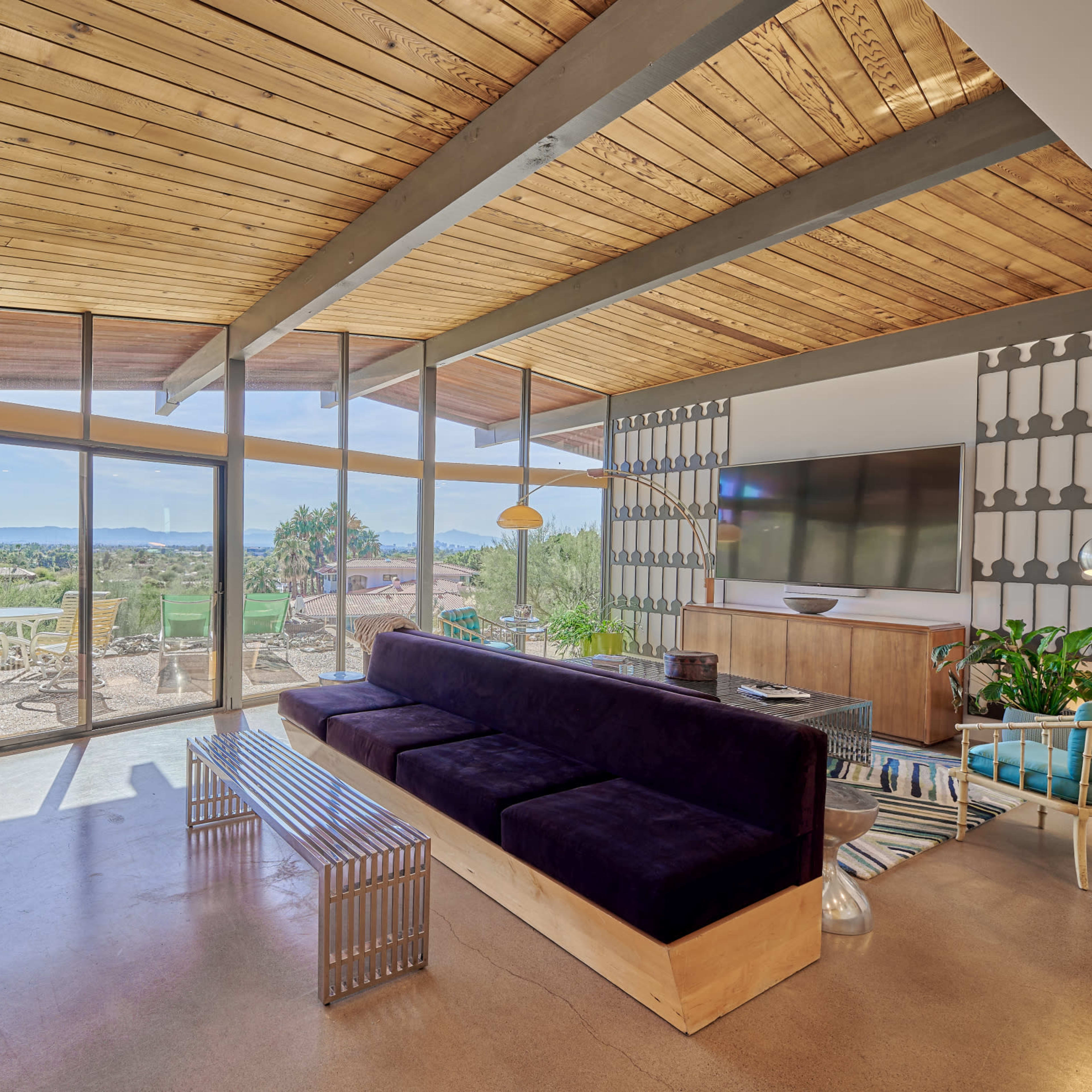 A modern living room features a large window with a view of the landscape, a dark sofa, a wooden bench, and a television on a wall unit.