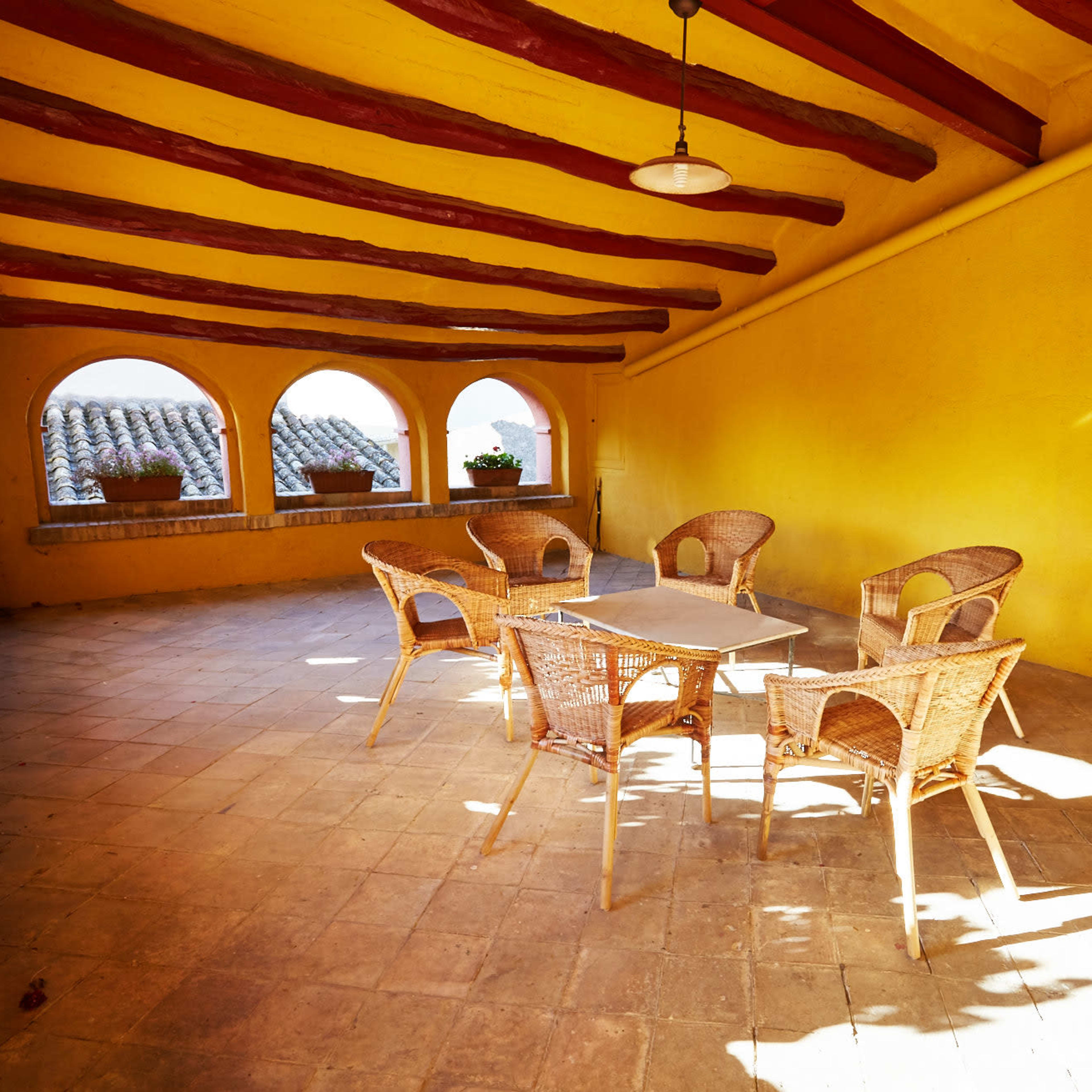 A sunlit patio with a table and four wicker chairs, surrounded by archways and potted plants.