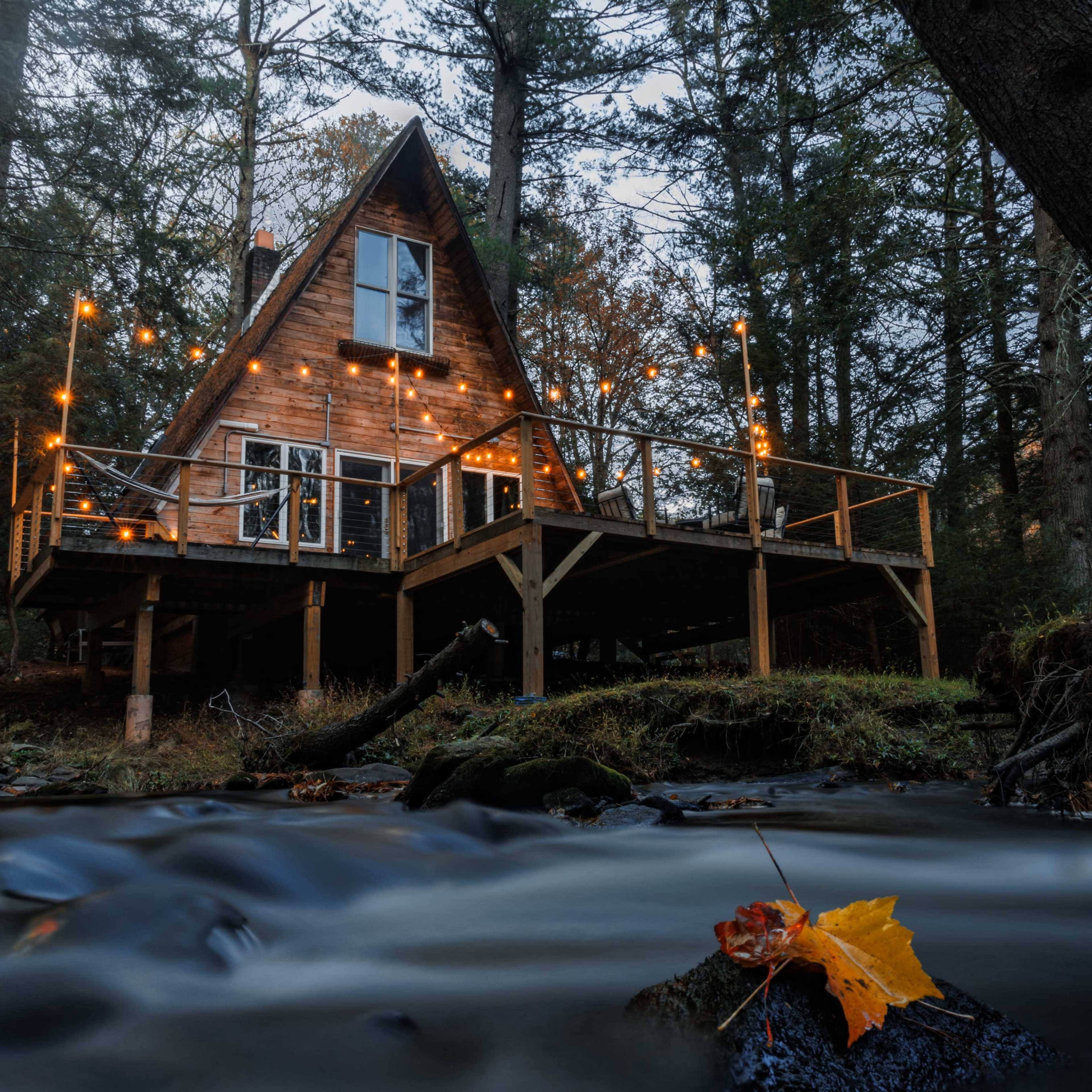 A wooden A-frame cabin with string lights sits on stilts by a flowing stream, surrounded by trees in a twilight setting.