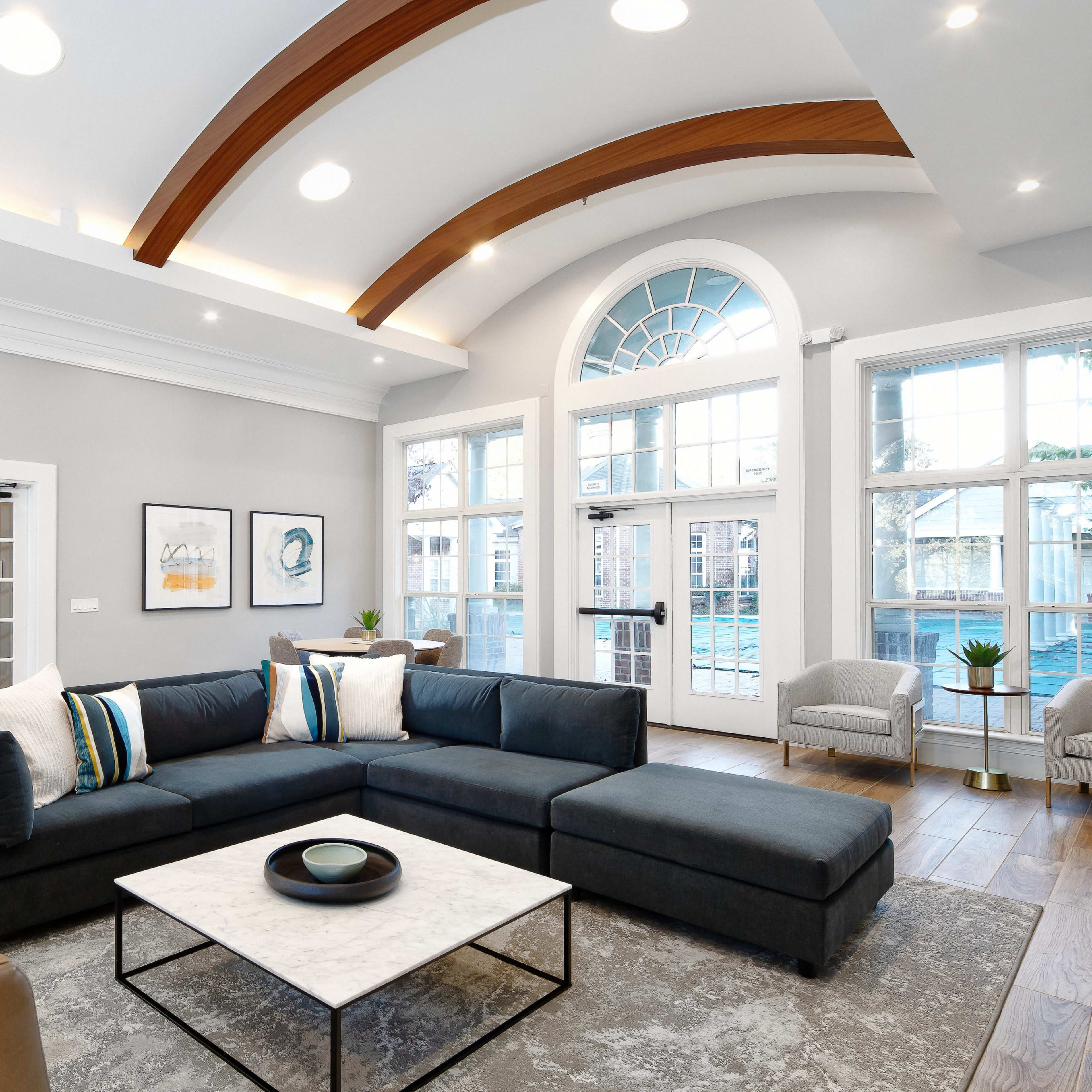A modern living area with a sectional sofa, a marble coffee table, and large windows allowing natural light to enter the space.