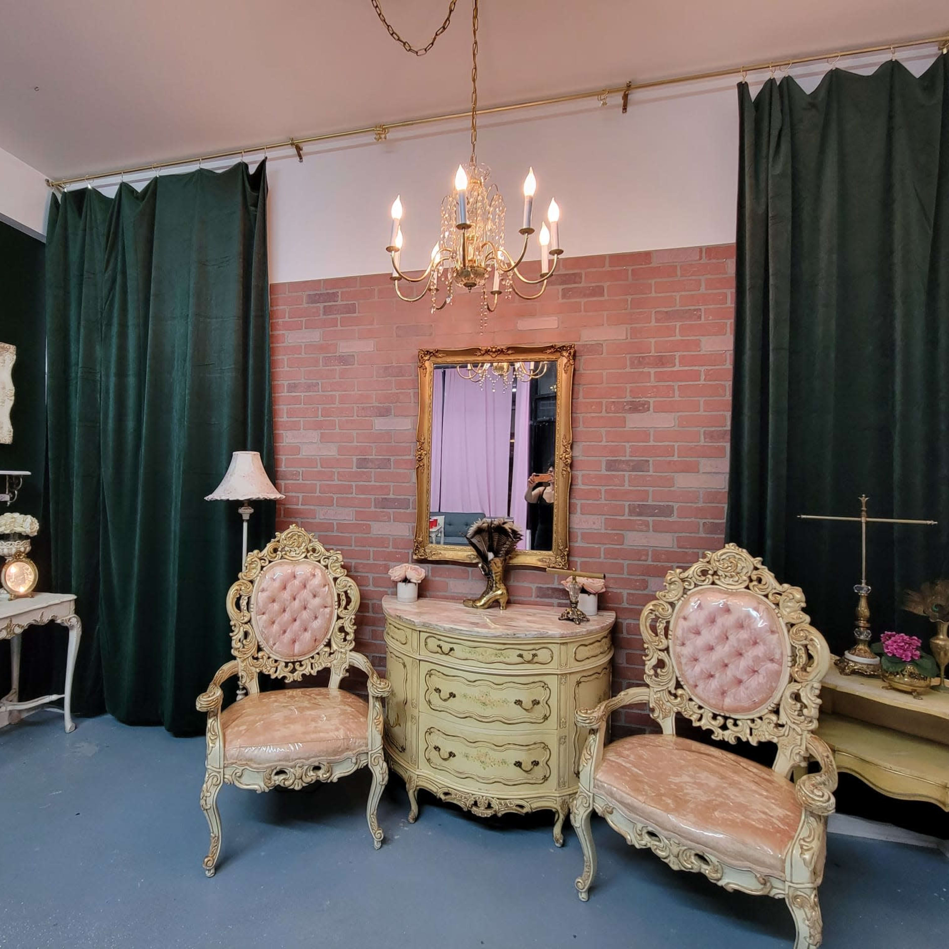 An elegantly arranged room with a vintage-style vanity set, two ornate chairs, a chandelier, and green curtains against a brick wall.