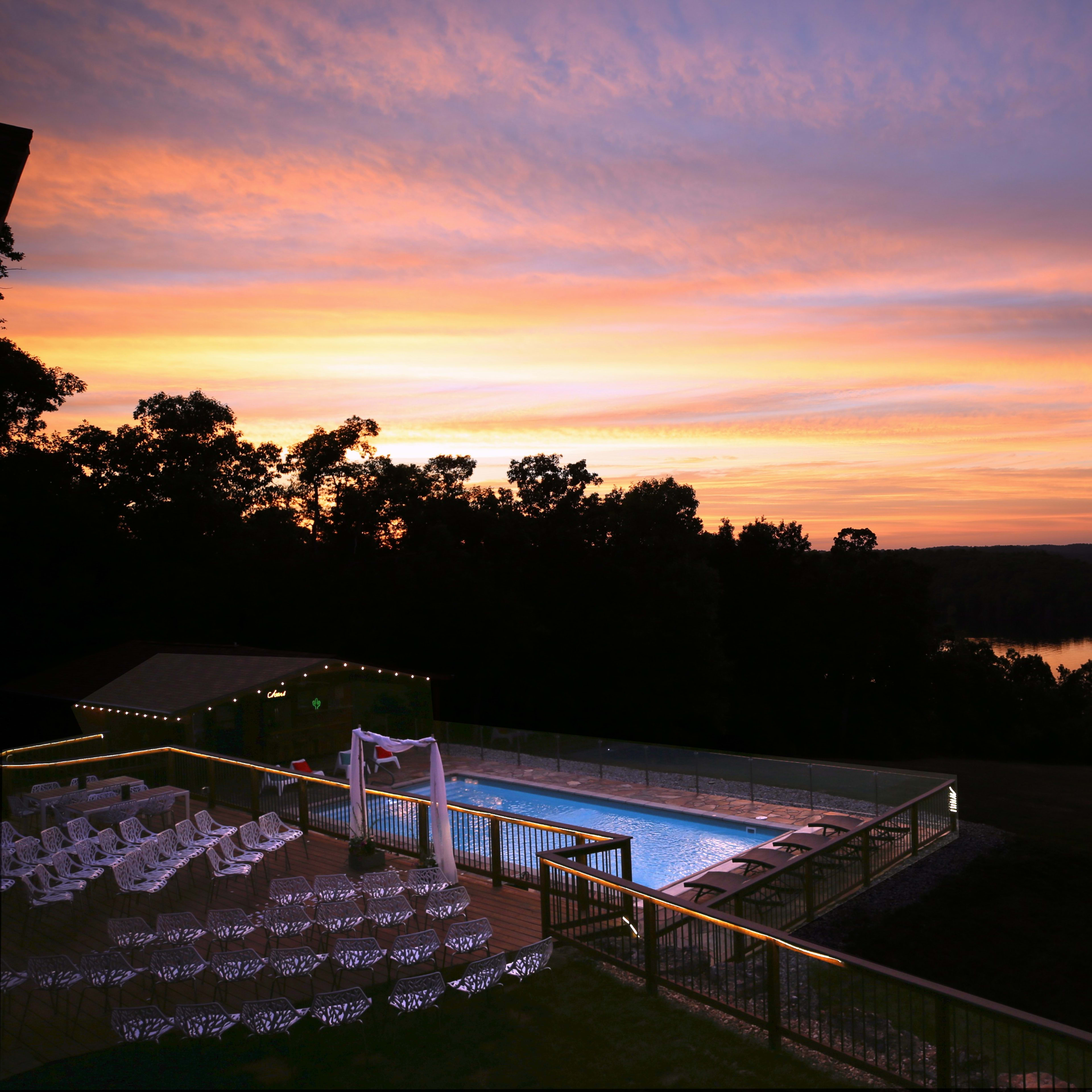 A swimming pool area with white chairs arranged around it, overlooking a lake at sunset.