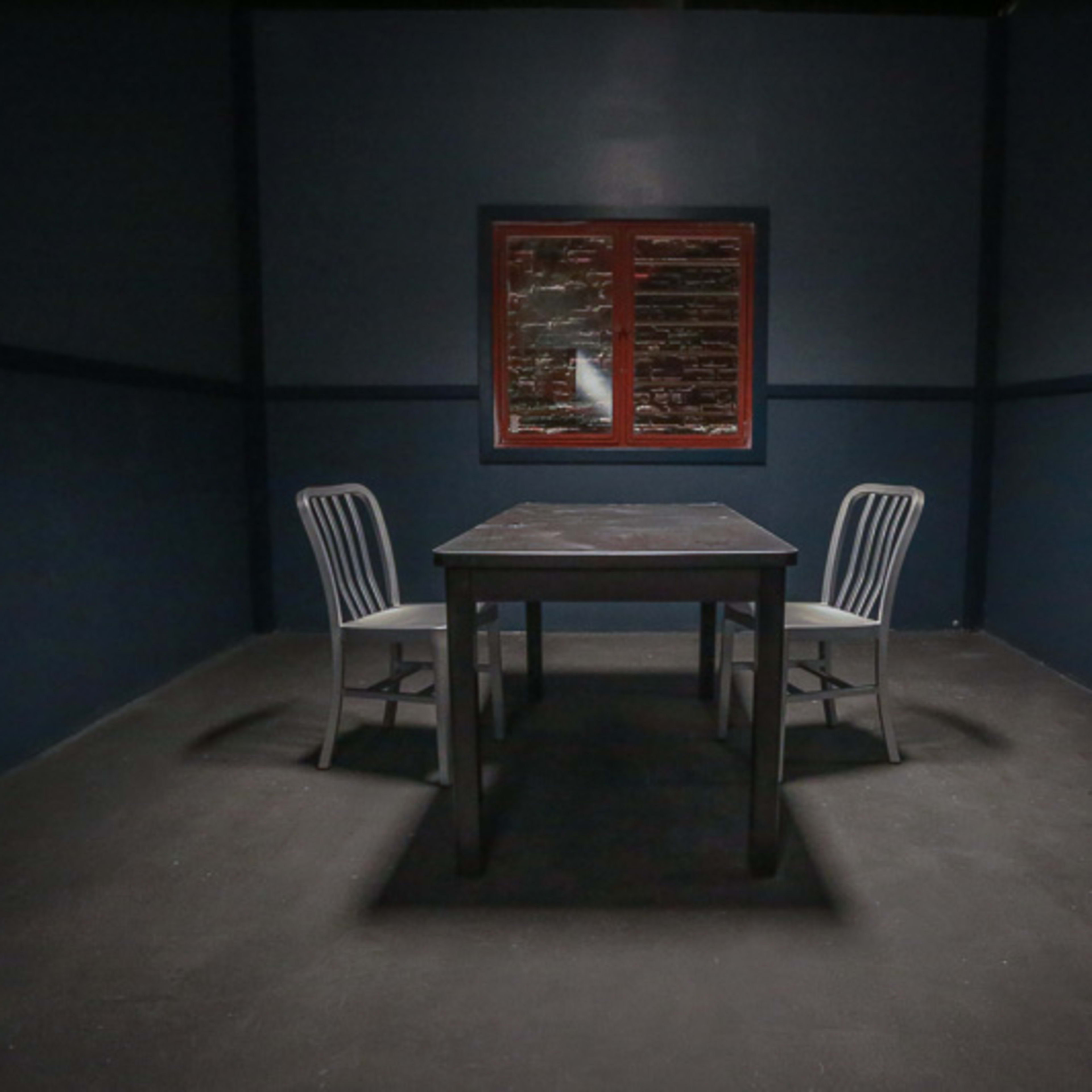 A dimly lit room features a table surrounded by two chairs and a window with a red frame.