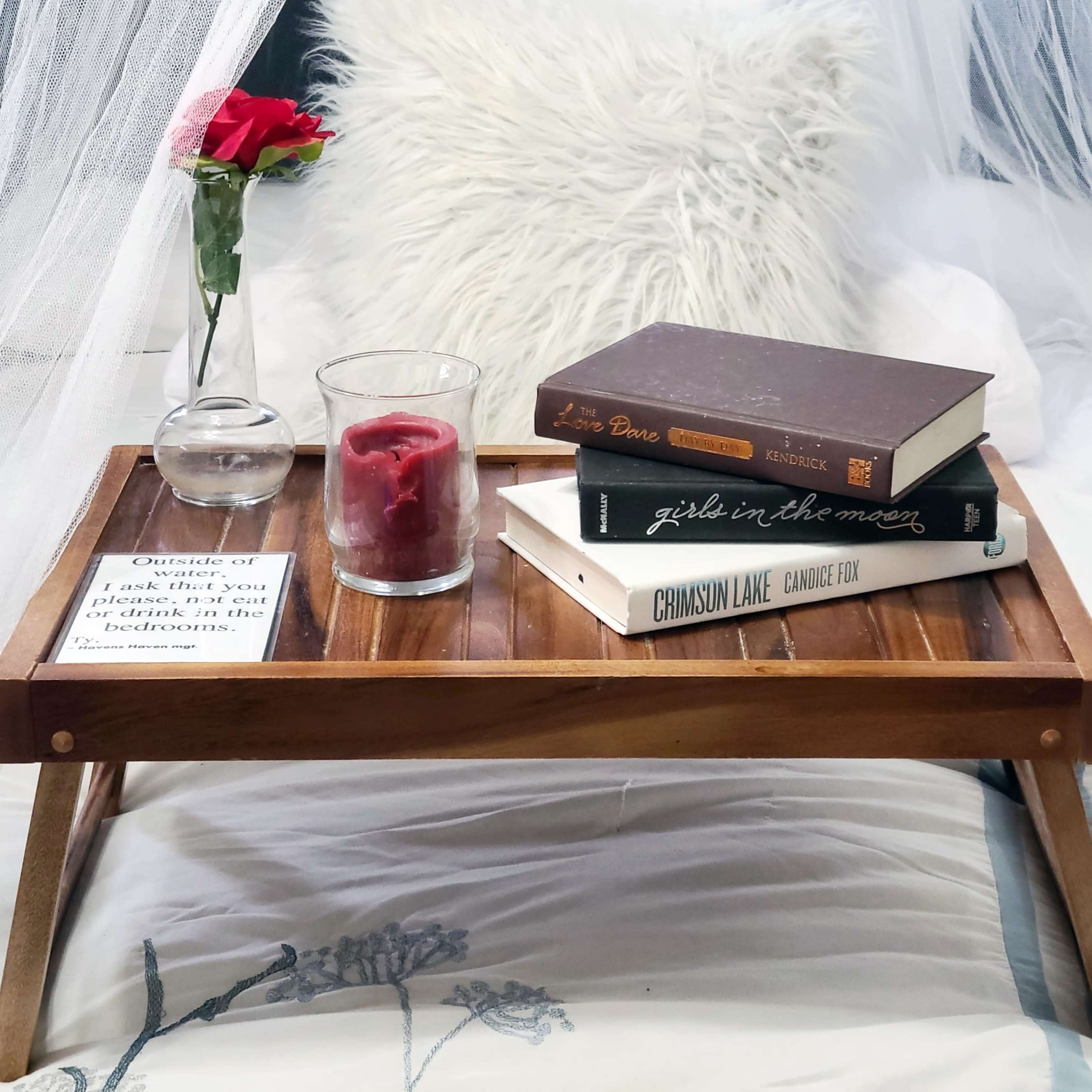 A wooden breakfast tray with a rose, a glass of water, a candle, and three books sits on a bed draped in a light canopy.