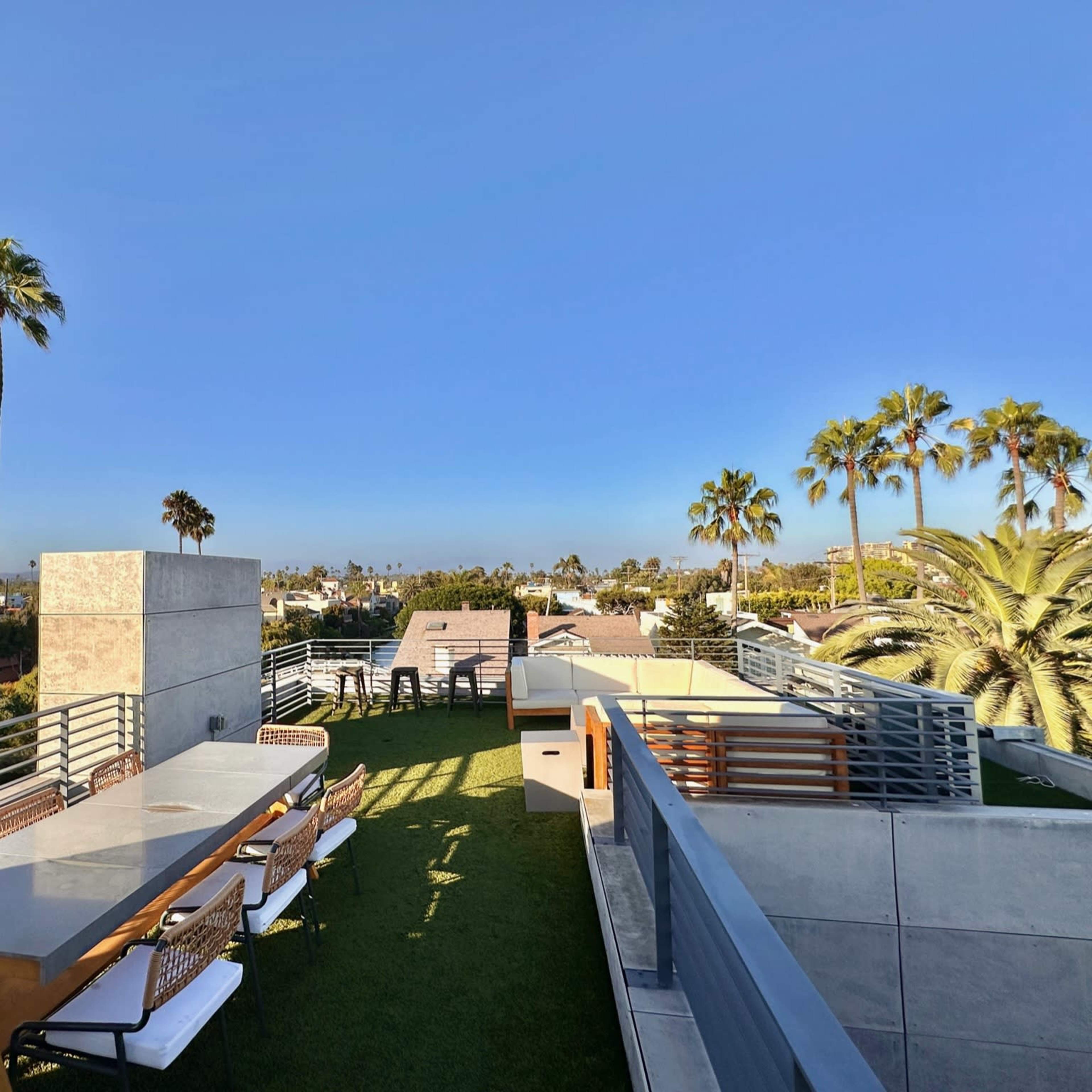 A rooftop terrace features a long table surrounded by chairs, with palm trees and a clear blue sky in the background.