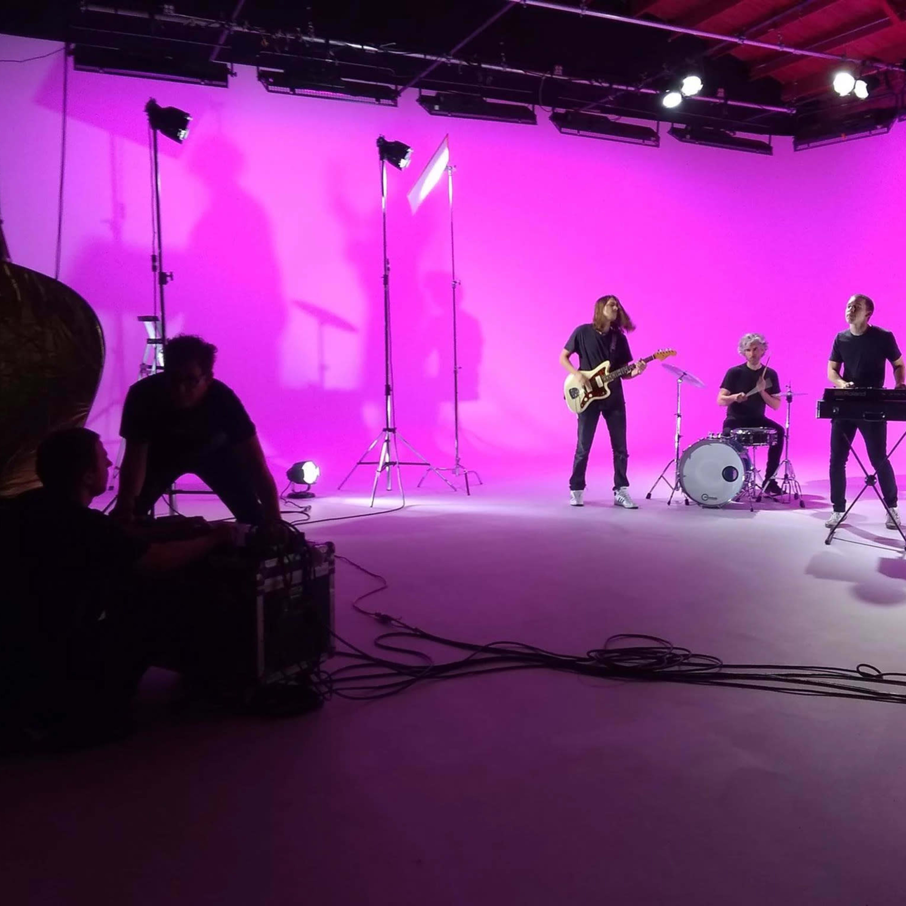 A band performs on a stage with a bright pink background, accompanied by lighting equipment and camera crew.