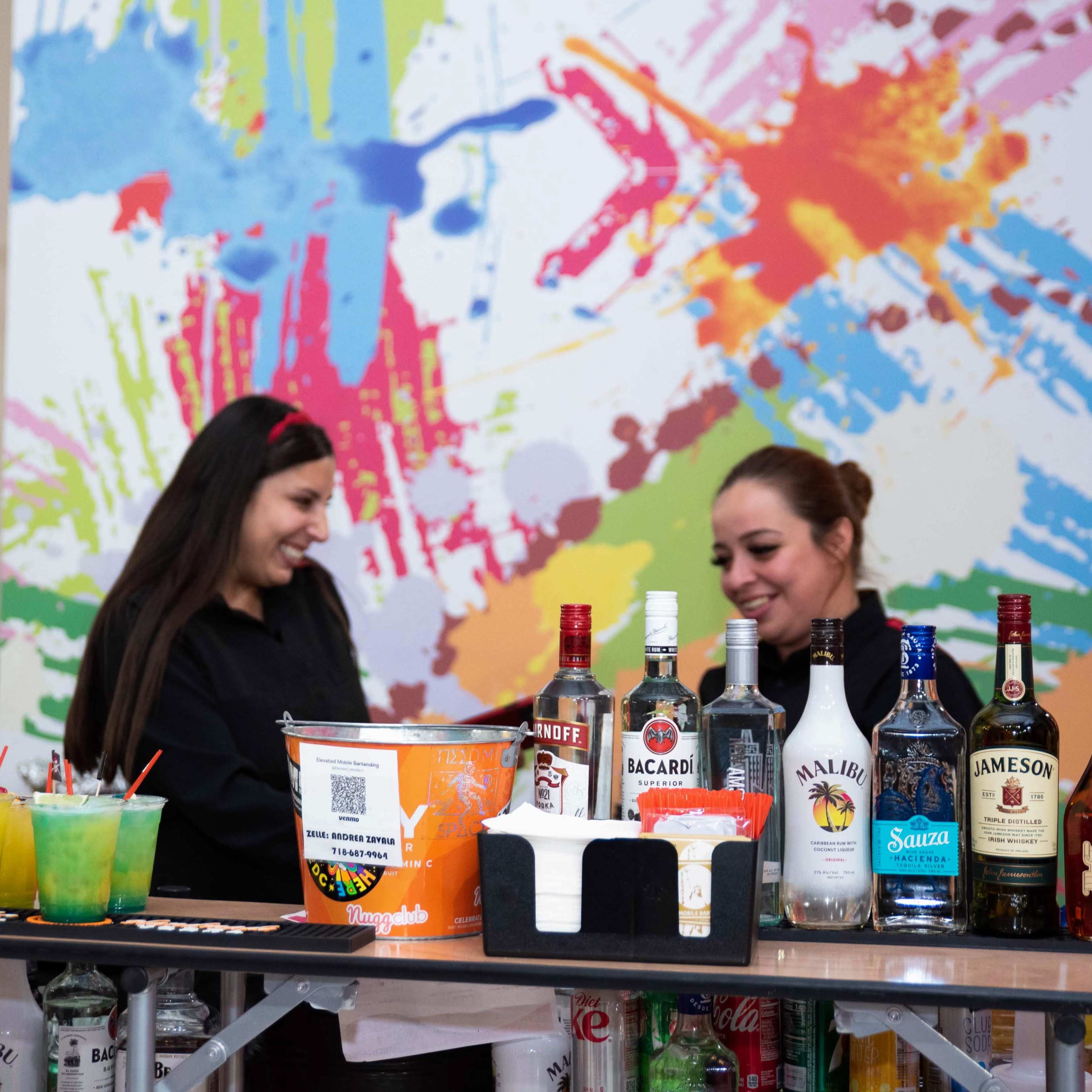 Two women stand behind a bar, interacting while surrounded by colorful splatter art and a variety of liquor bottles.