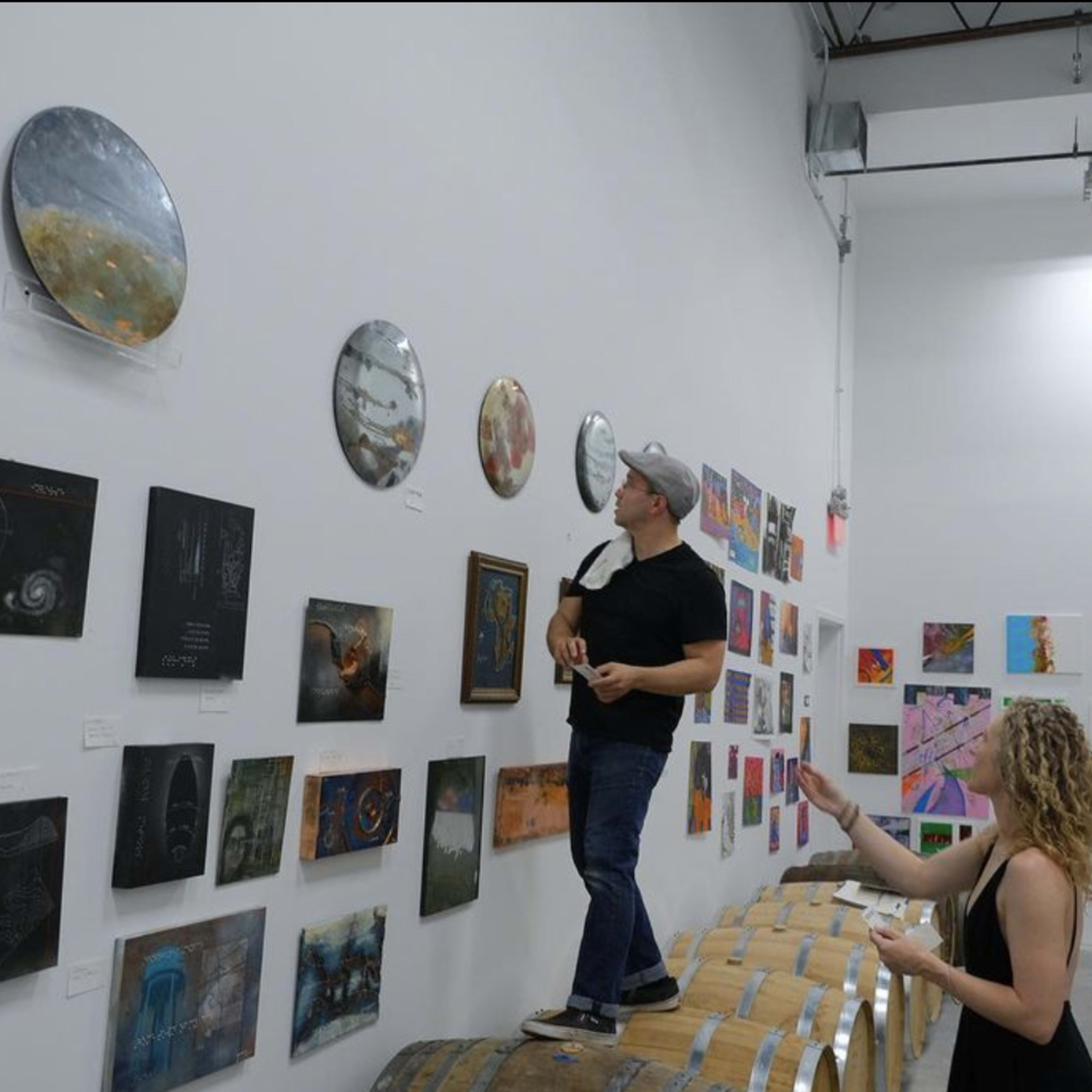 A man stands on a barrel while inspecting art pieces on a wall in a gallery, as three other individuals observe nearby.