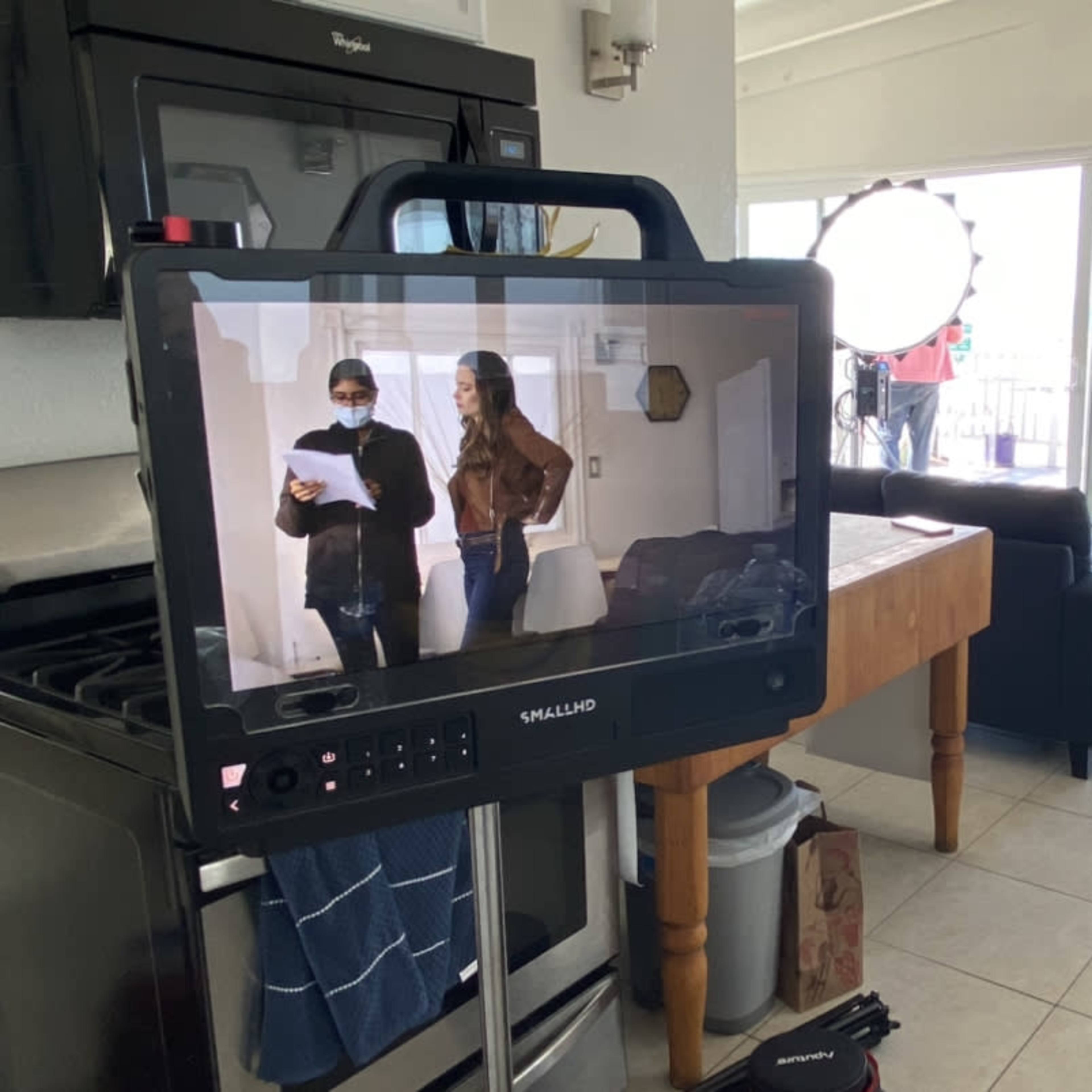 A filming crew sets up a scene in a bright room with a monitor displaying two actors rehearsing their lines.