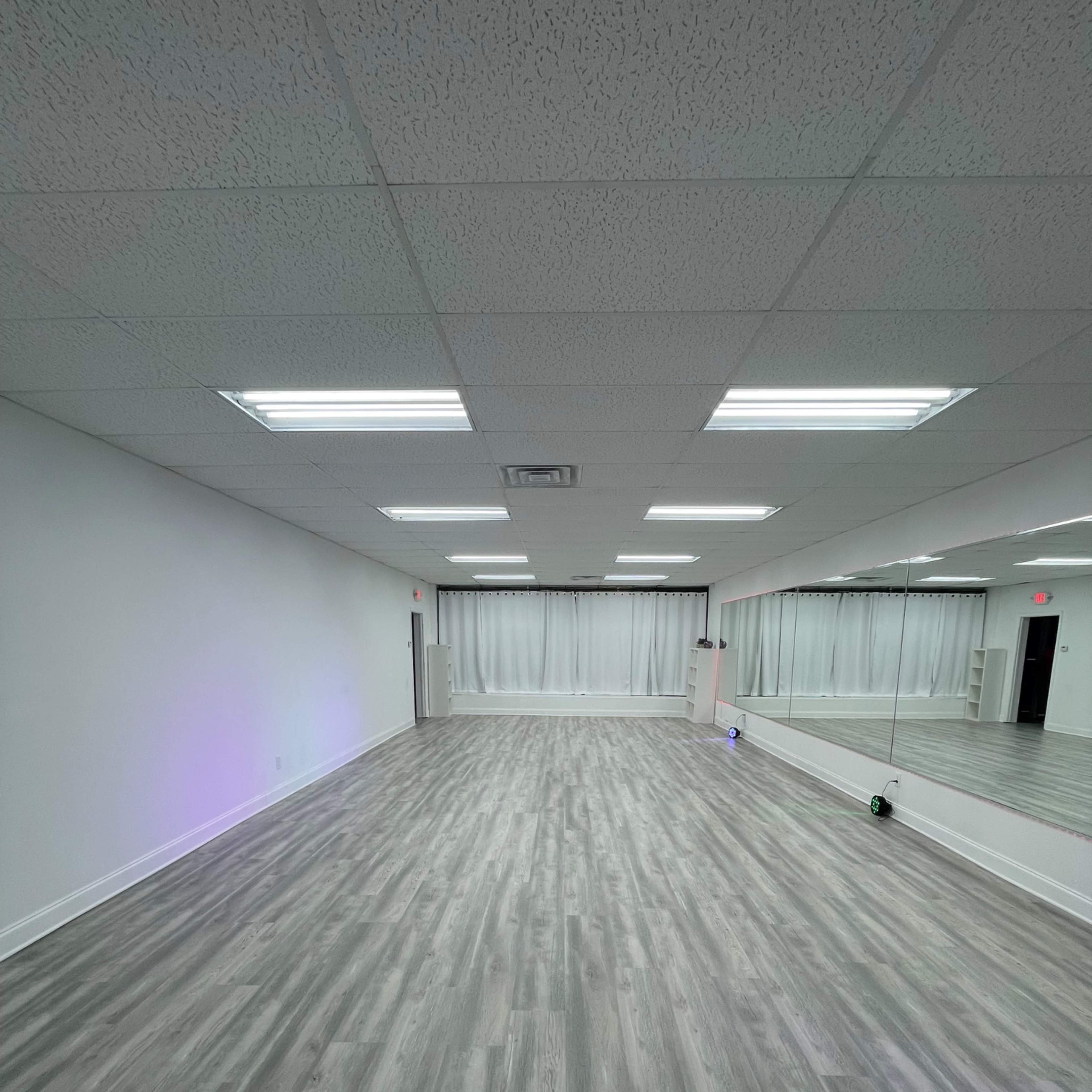 An empty room with mirrored walls, light-colored wood flooring, and fluorescent ceiling lights.