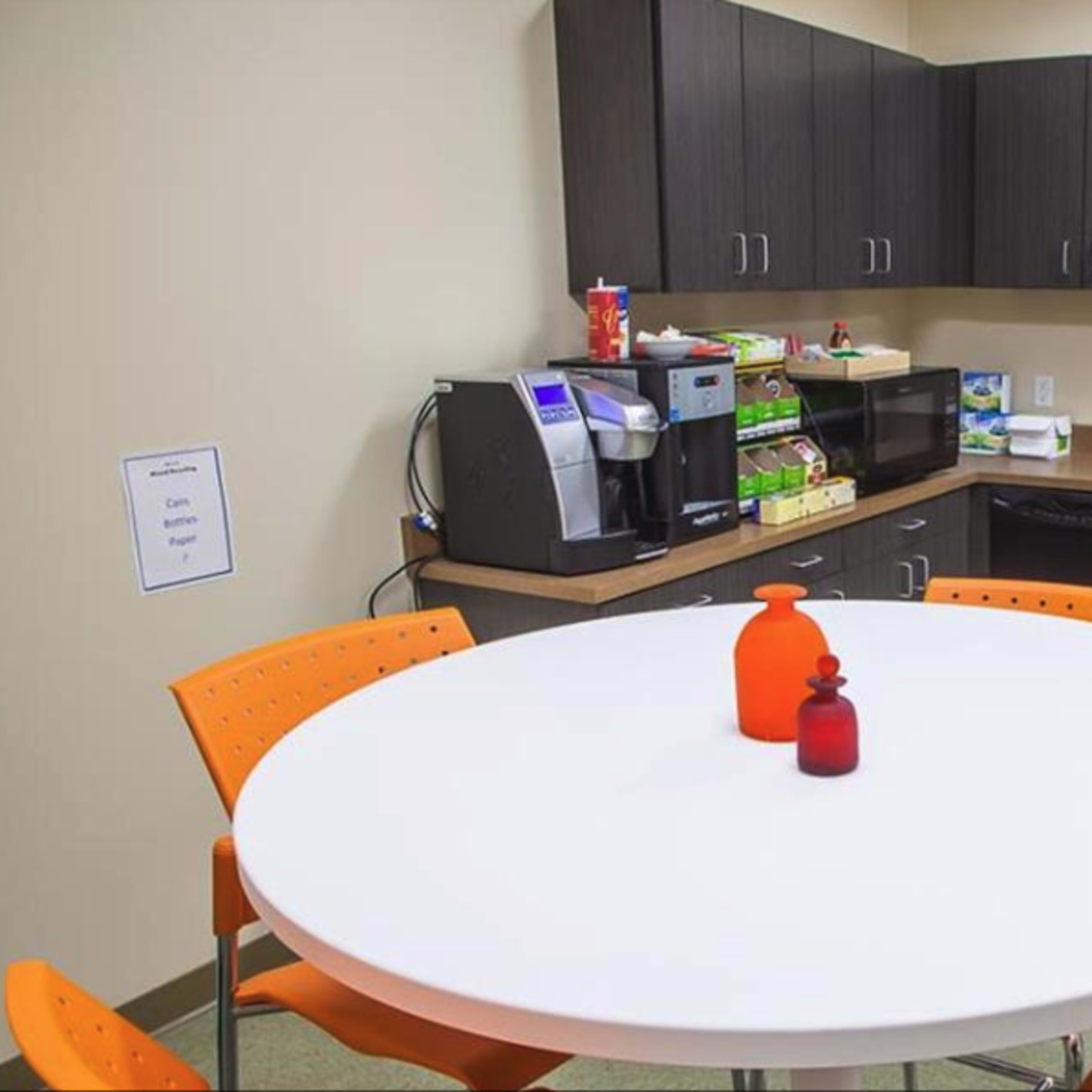 A break room features a round white table surrounded by orange chairs, with kitchen cabinets, a coffee machine, and a black refrigerator in the background.