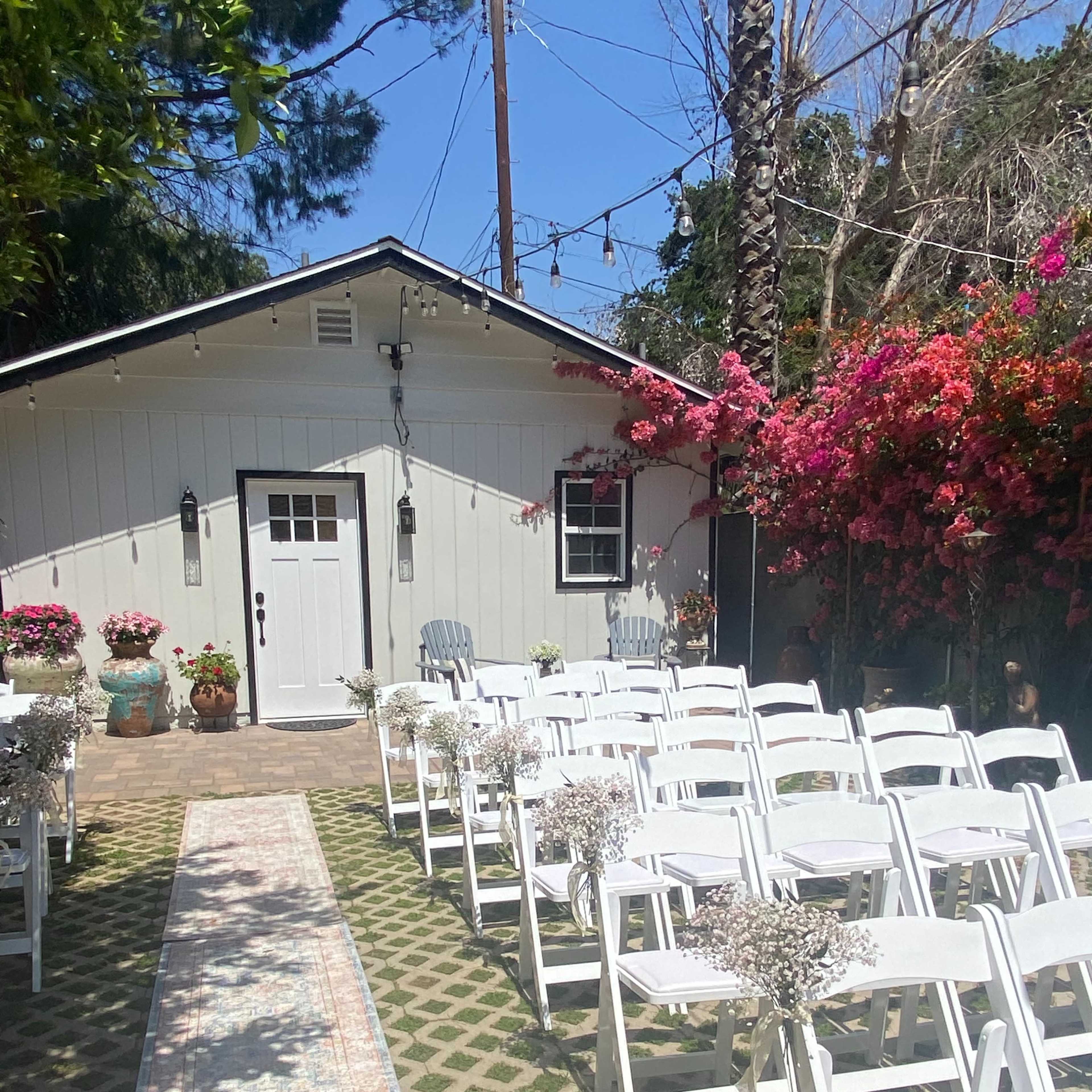 A path lined with white chairs leads to a small white building surrounded by colorful flowers and greenery.