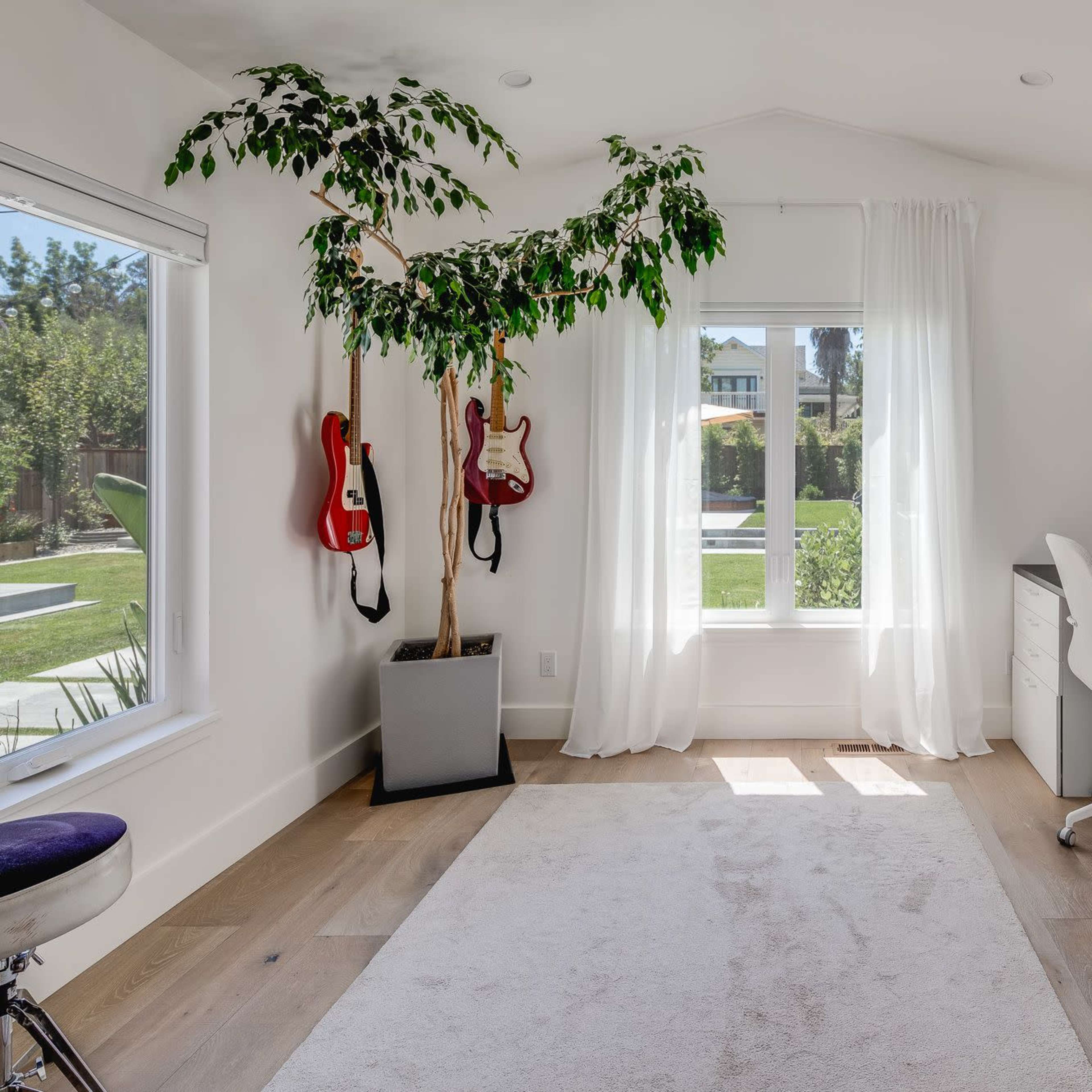 A modern home office with two desks, a potted plant, and guitars hanging on the wall beside a large window allowing natural light.