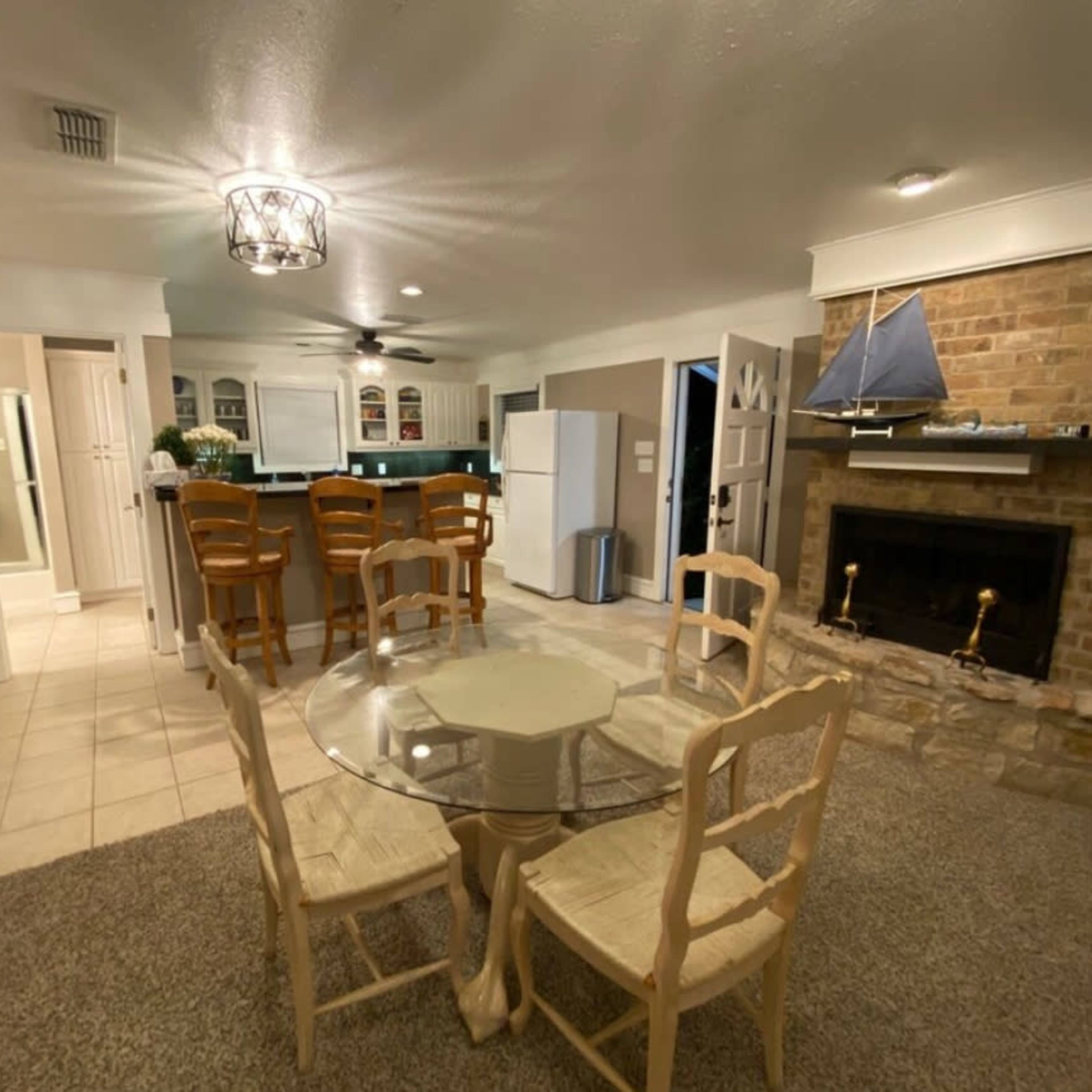 A light-colored kitchen and dining area with a round glass table, four chairs, a stone fireplace, and a bar with high stools.