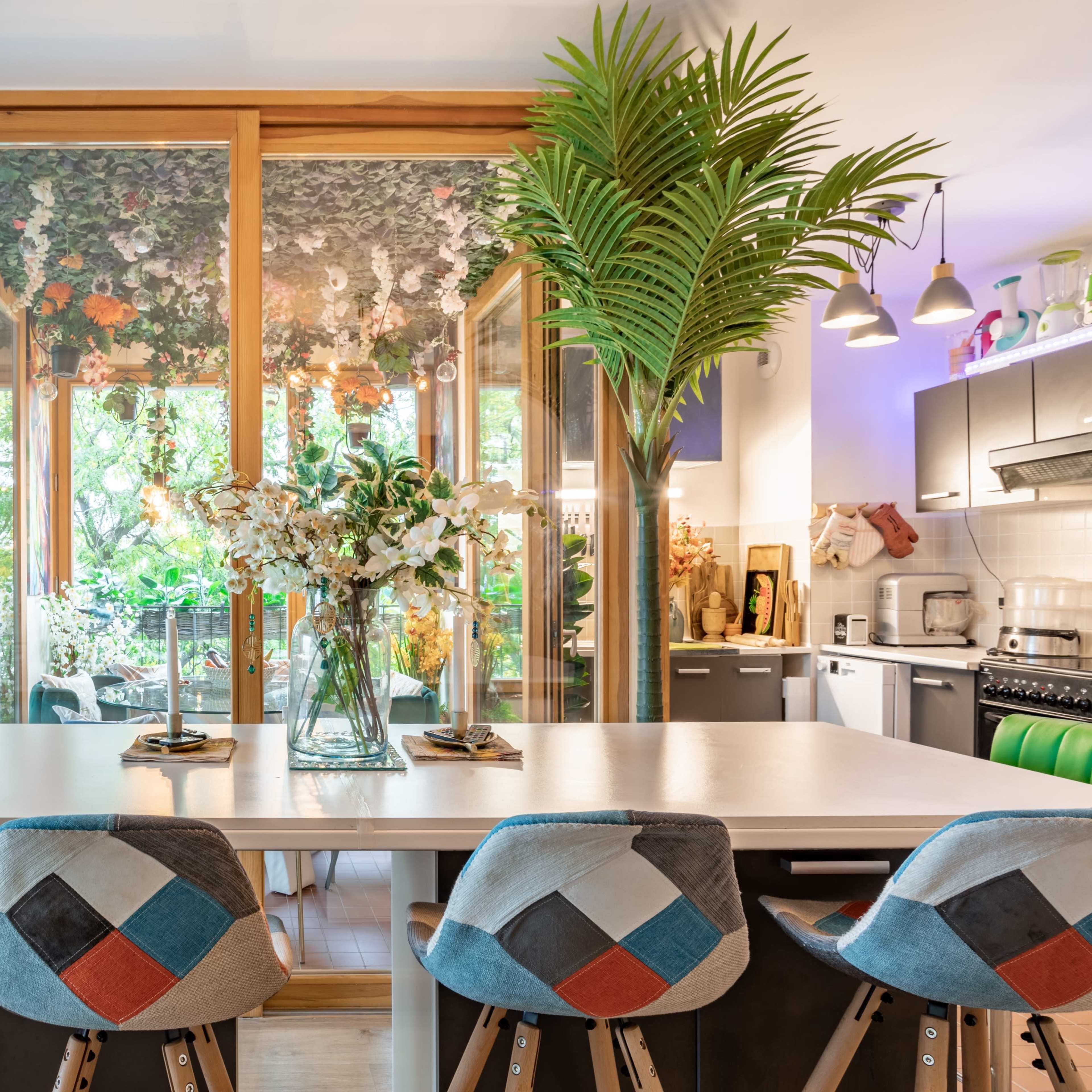 A modern kitchen with a dining area featuring a large white table, colorful upholstered bar stools, and a glass-enclosed room filled with plants and flowers.